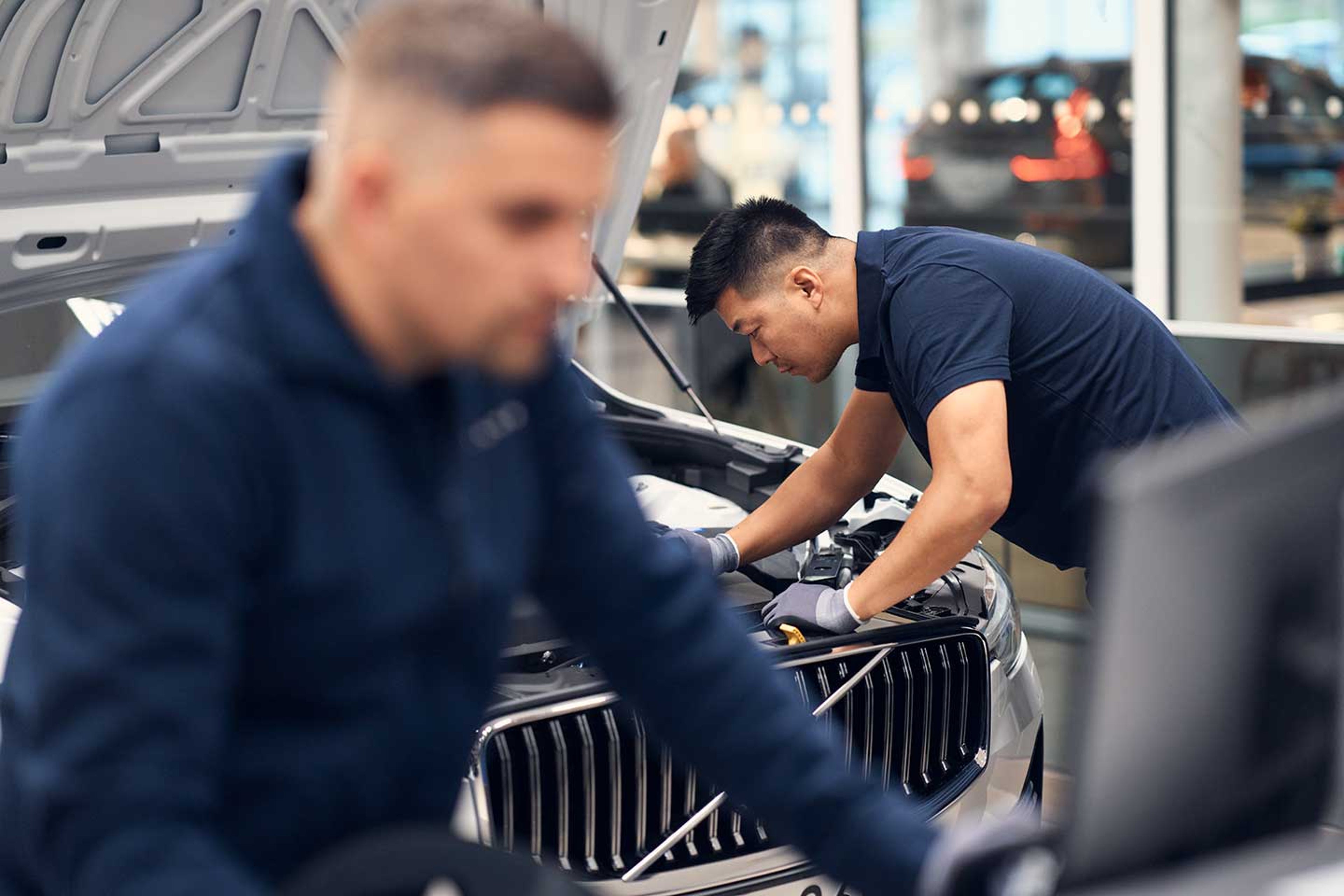 To mænd og fronten på en bil fra Volvo med kølerhjelmen åben ses, der er fokus på den bagerste person der kigger ned i motoren på bilen, mens den anden arbejder på en computer.