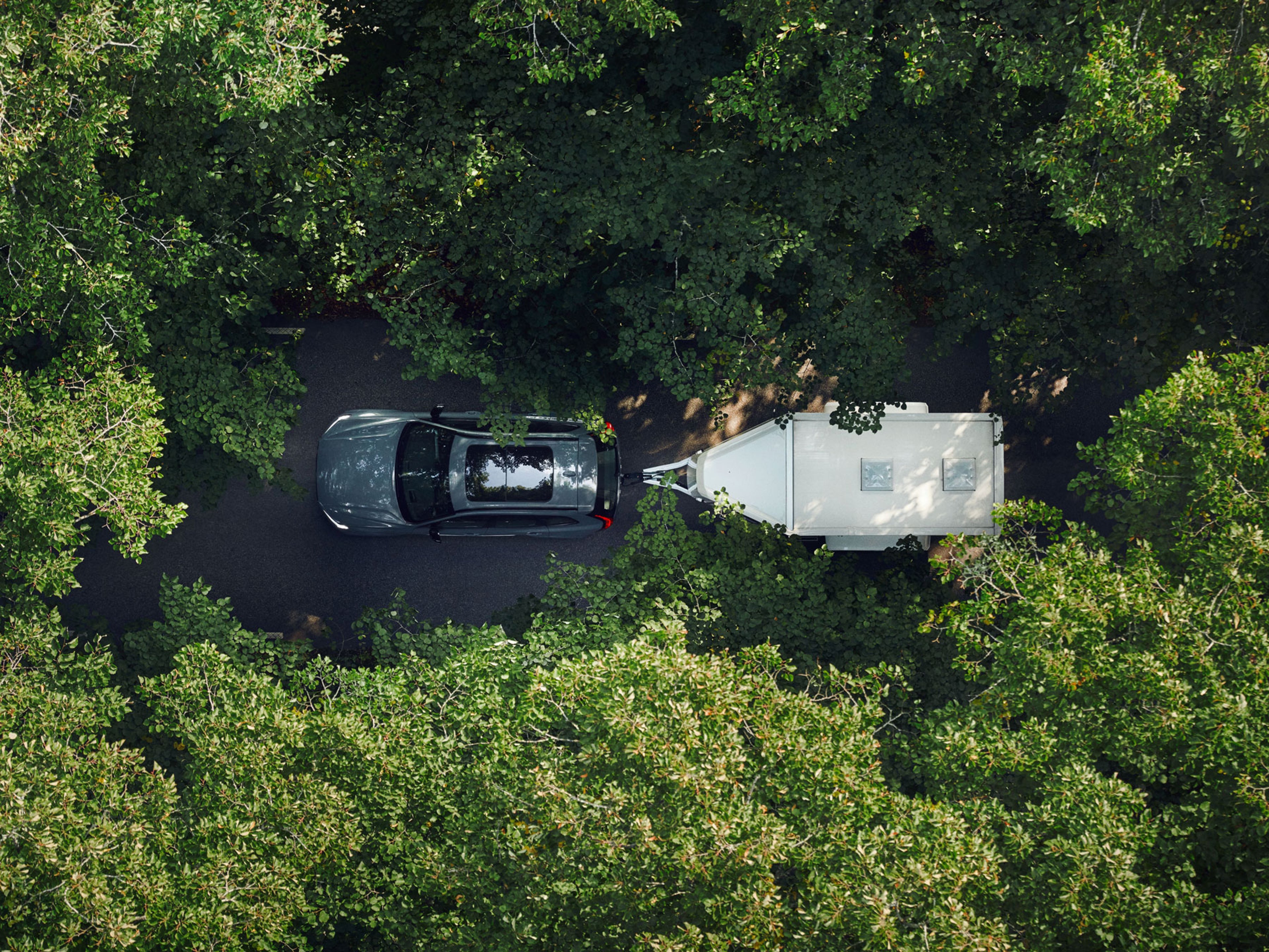 Vue aérienne d'une voiture tractant une remorque sur une route étroite entourée d'arbres verts