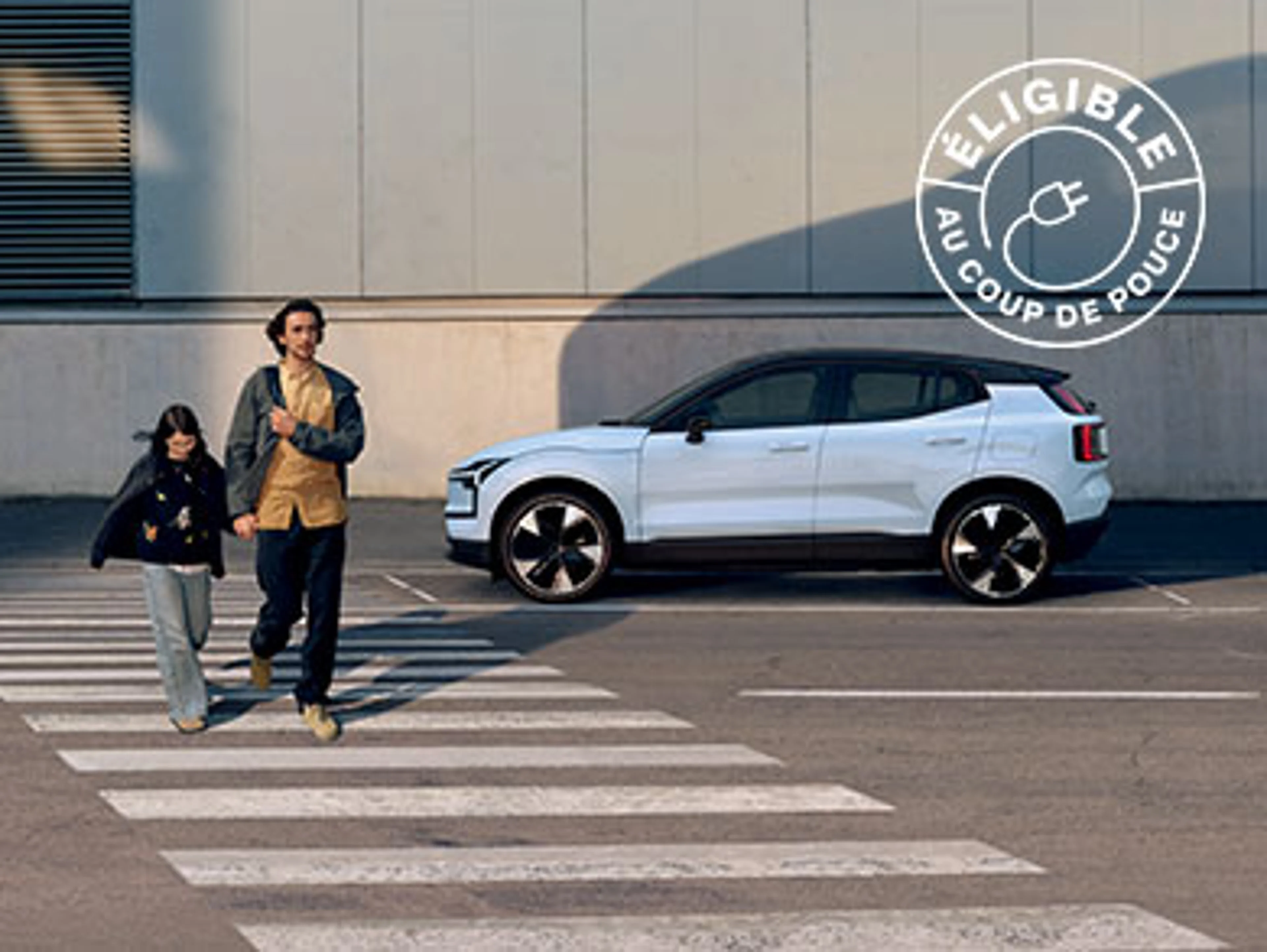 Deux personnes traversent un passage piéton devant une voiture Volvo EX30 blanche et un bâtiment industriel.