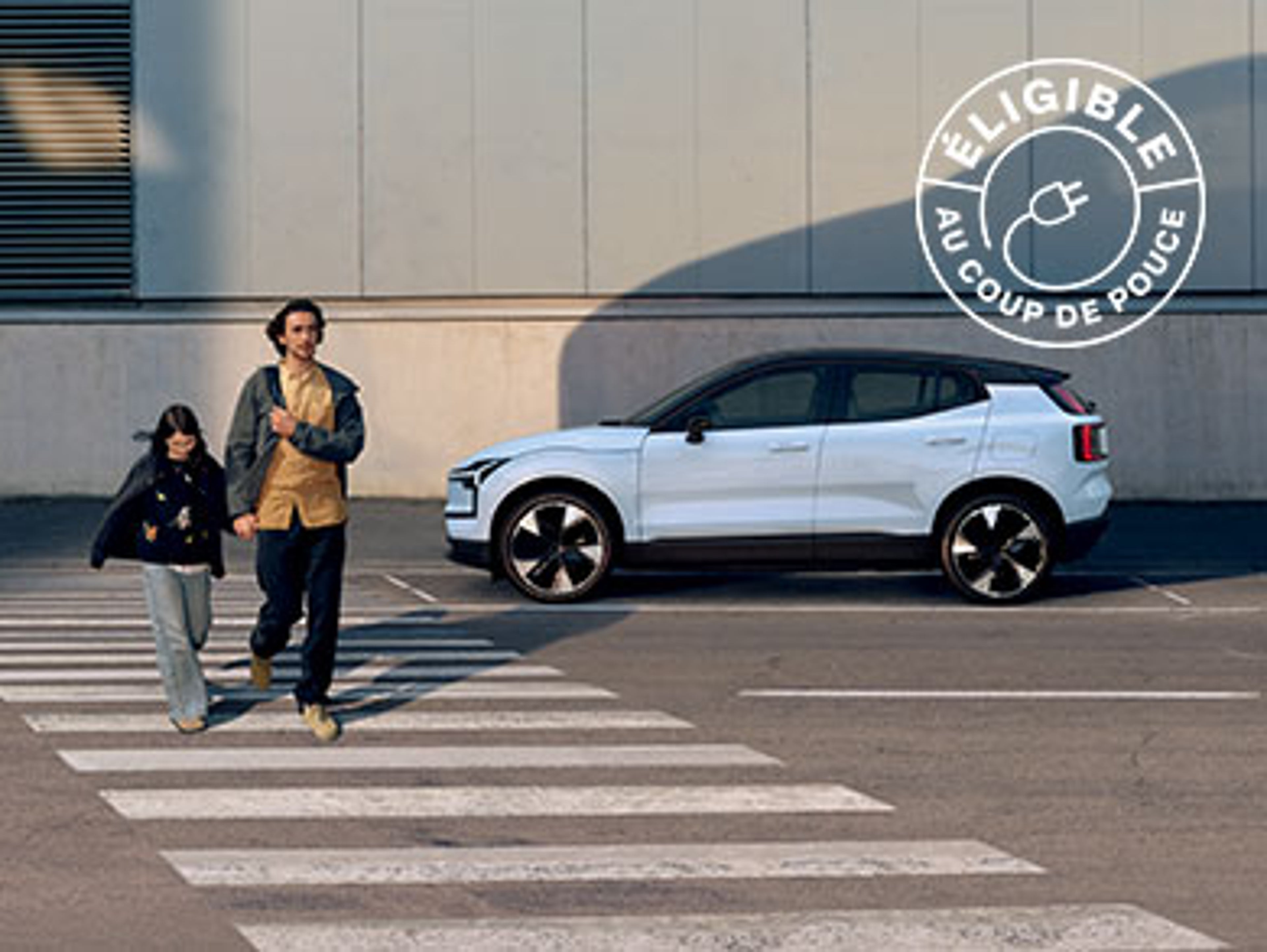 Deux personnes traversent un passage piéton devant une voiture Volvo EX30 blanche et un bâtiment industriel.