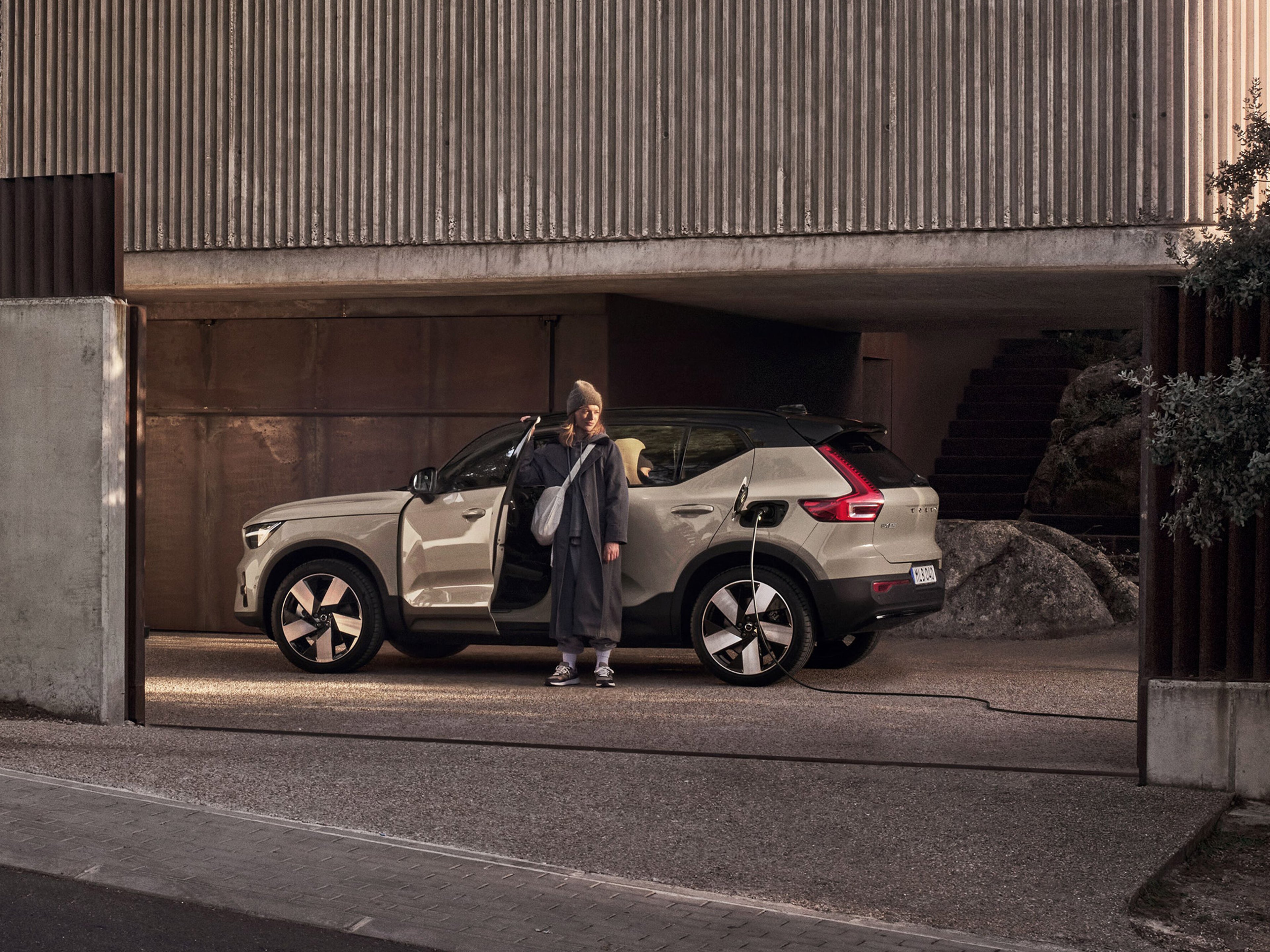 Nainen seisoo pihalla lataukseen kytketyn Sand Dune -värisen Volvo EX40 täyssähköauton edessä.