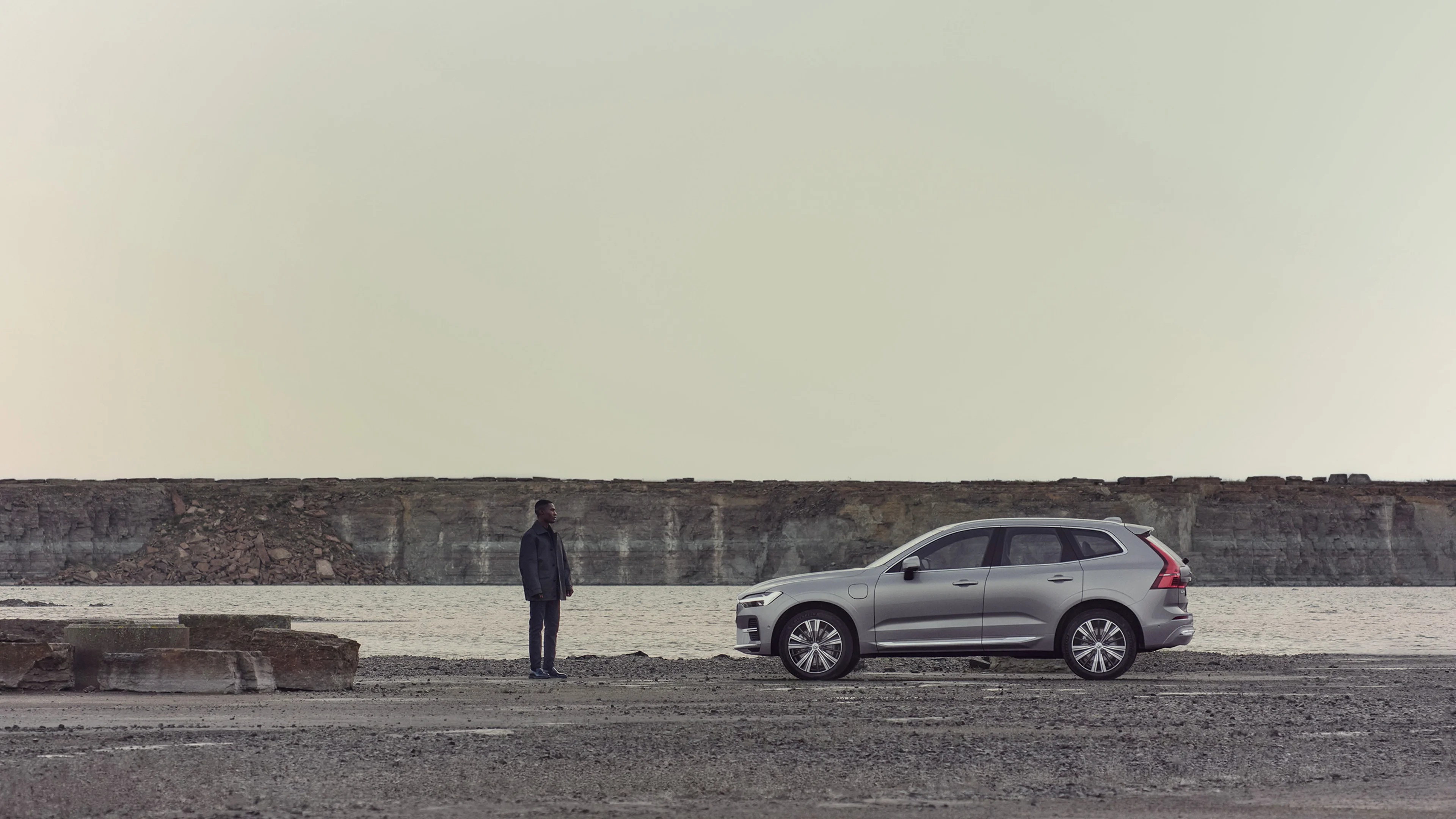 Ein Mann steht vor einem silbernen Volvo XC60 SUV, der auf einer Schotterstraße nahe eines Sees steht.