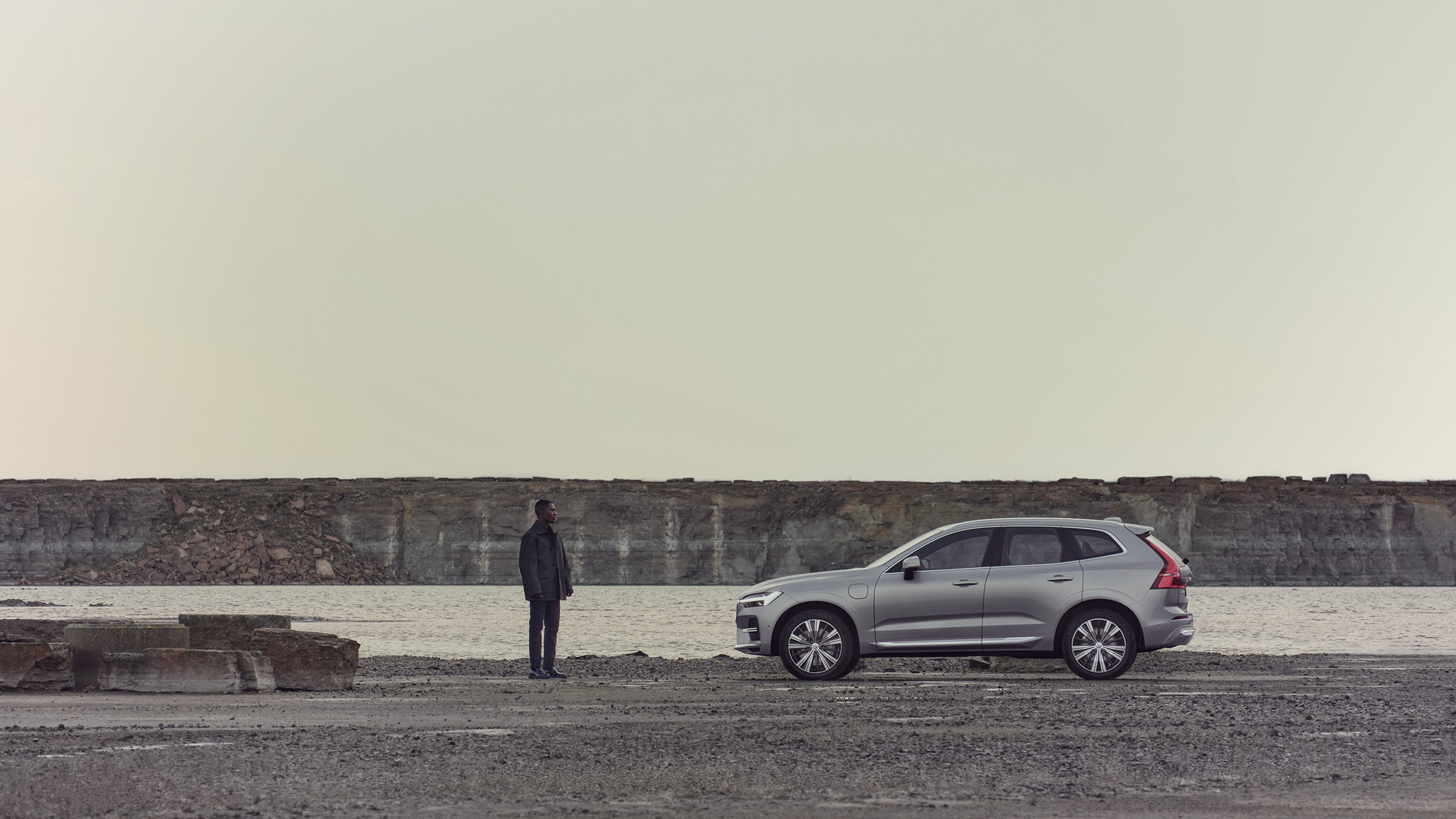Ein Mann steht vor einem silbernen Volvo XC60 SUV, der auf einer Schotterstraße nahe eines Sees steht.