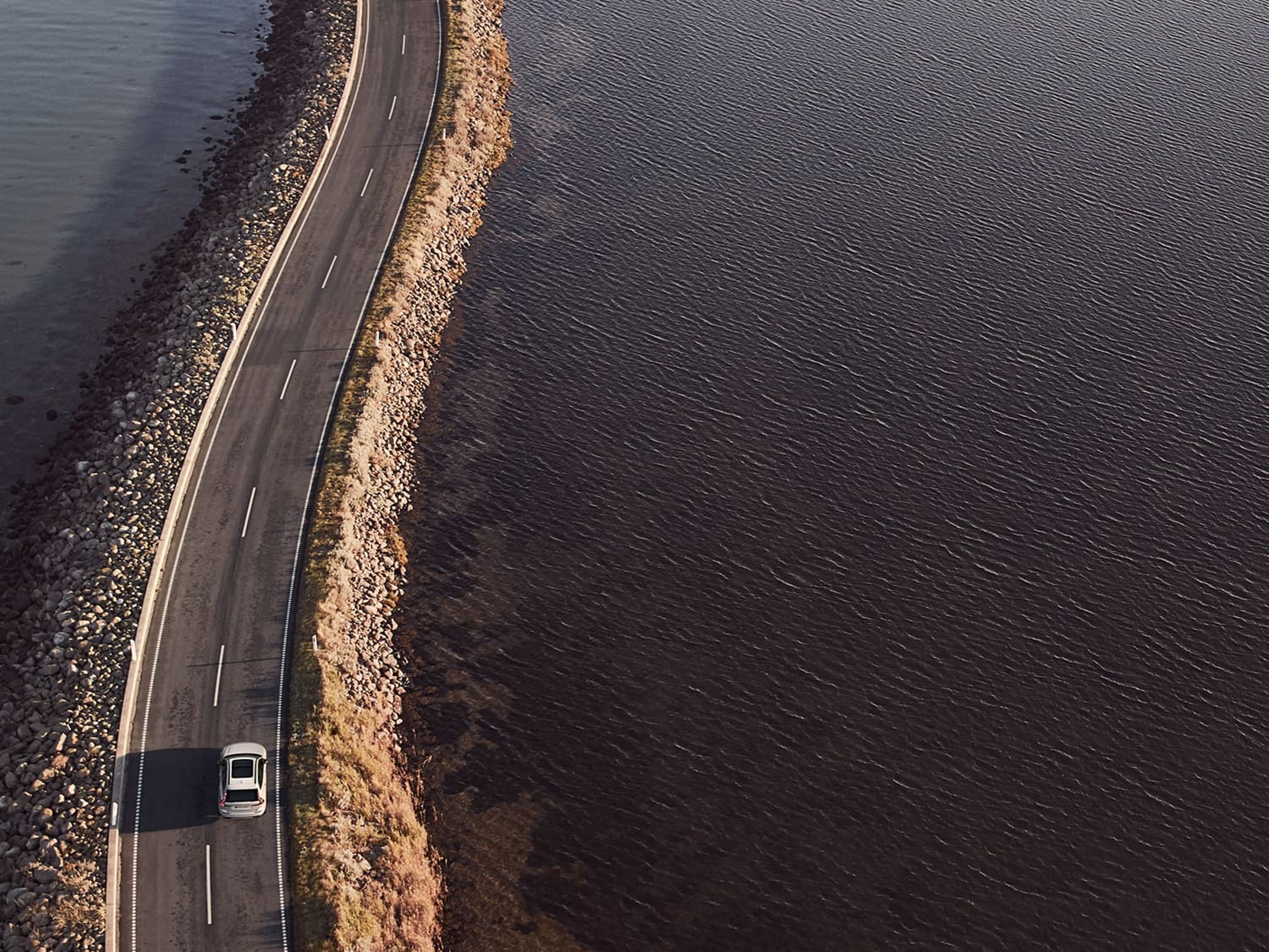 Vue aérienne d'une Volvo blanche roulant sur une route longeant l'eau, entourée d'un littoral rocheux.