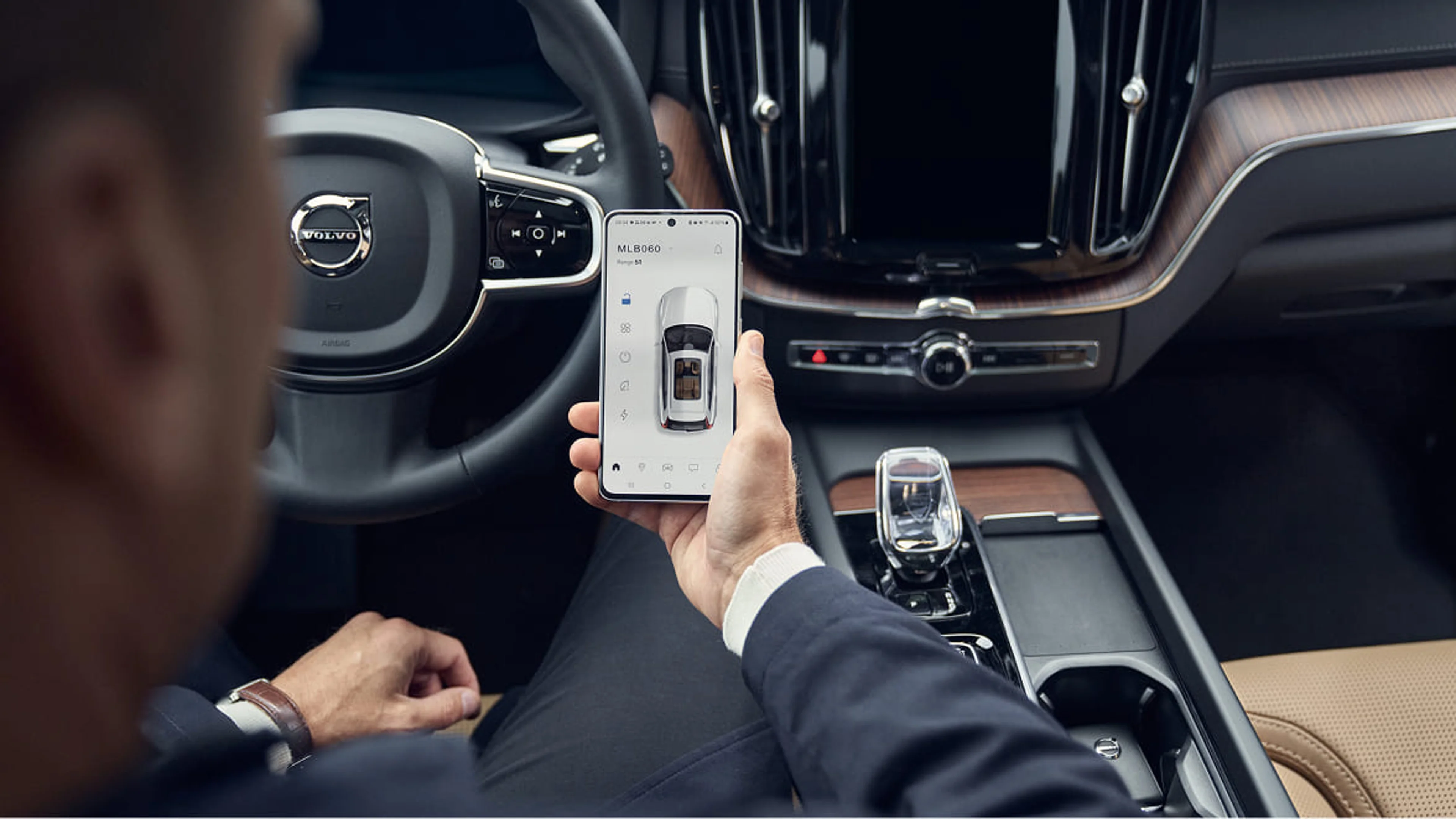 Hand som håller i en telefon med Volvo Cars app. Ratt och växelspak i bakgrunden.