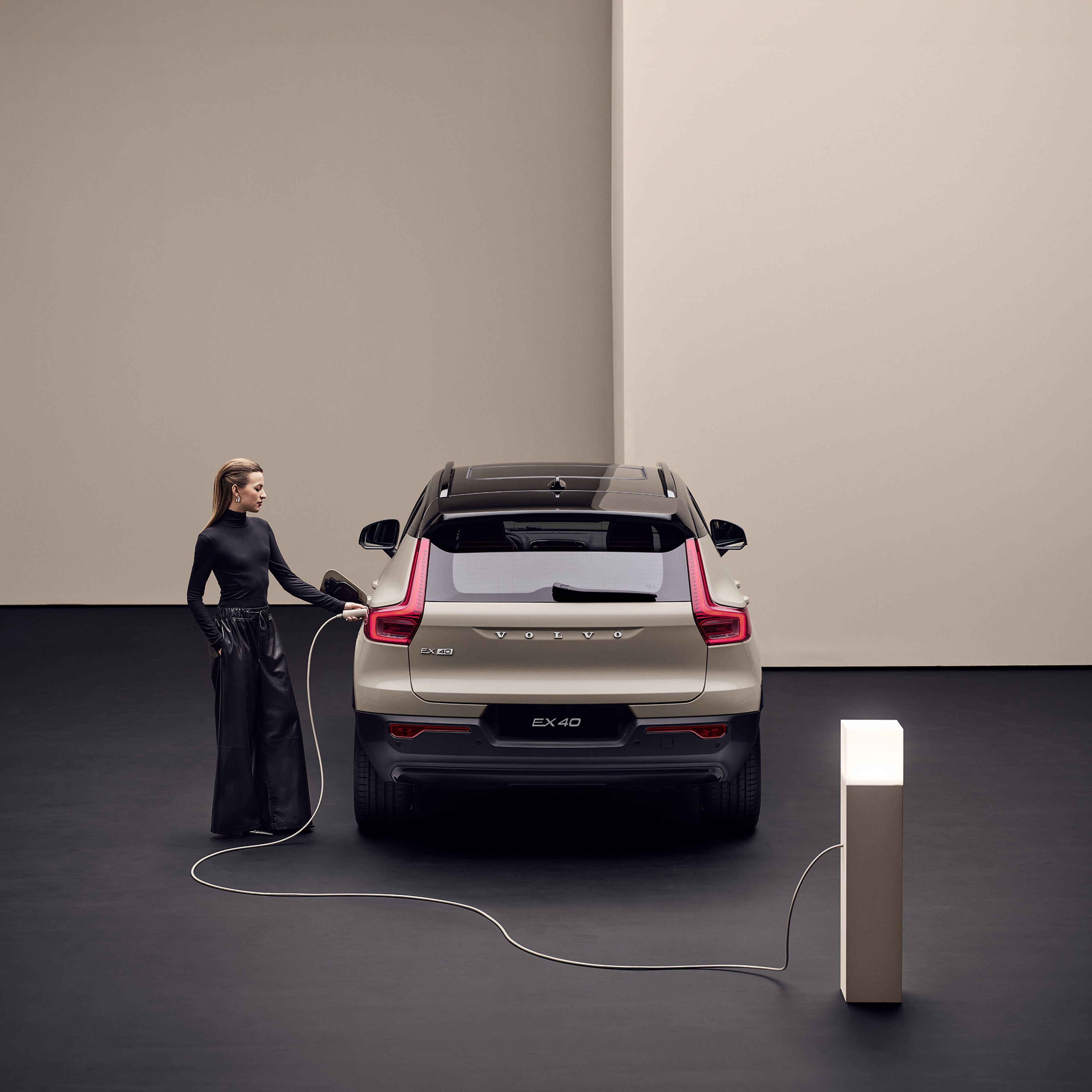 A woman in black clothing charging a sand dune fully electric Volvo EX40 from the rear at a modern indoor charging station, with a minimalist background.