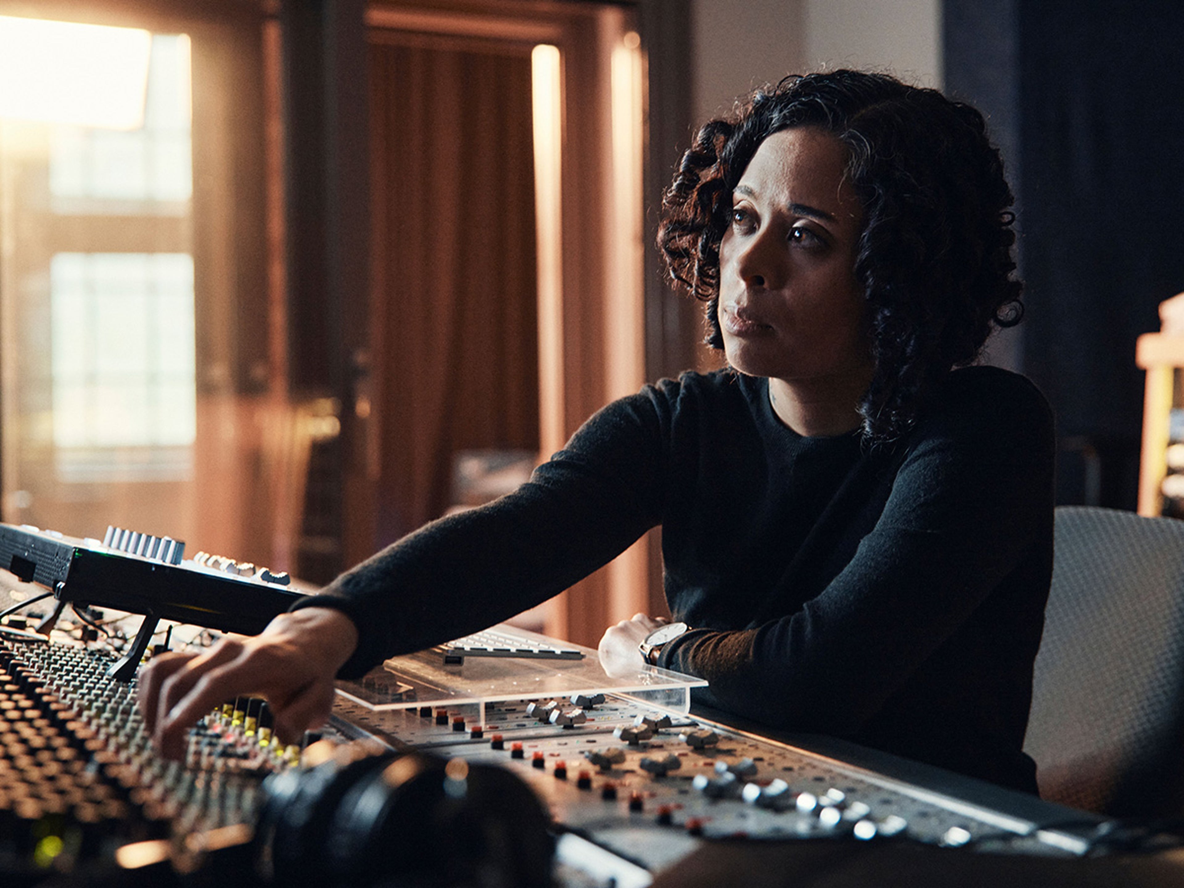 Een vrouw met donker krullend haar zit achter een mengpaneel in een opnamestudio.