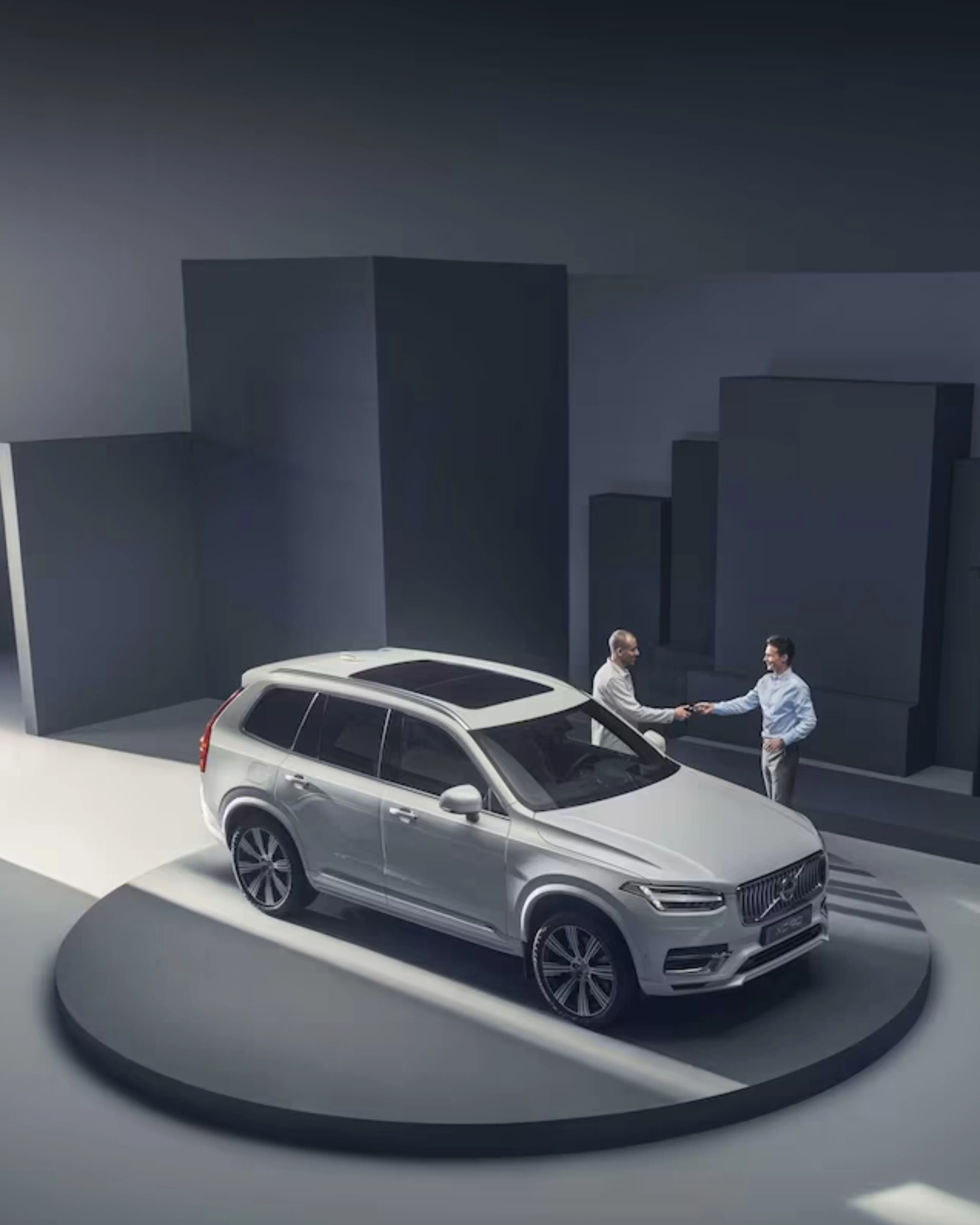 Ein grauer Volvo steht auf einer etwas erhöhten Plattform in einem Studio. Dahinter stehen zwei Menschen, die sich die Hand schütteln.