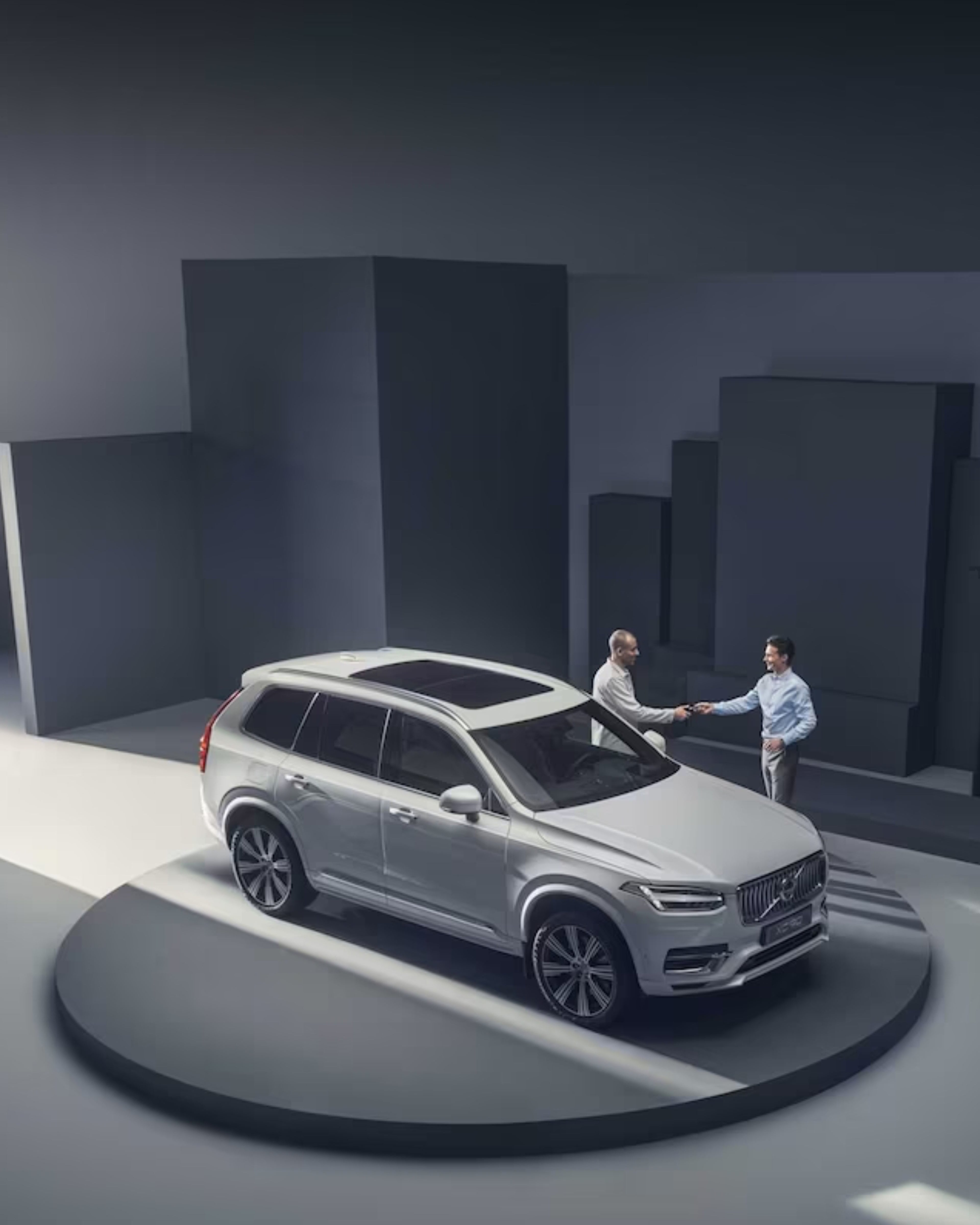 Ein grauer Volvo steht auf einer etwas erhöhten Plattform in einem Studio. Dahinter stehen zwei Menschen, die sich die Hand schütteln.