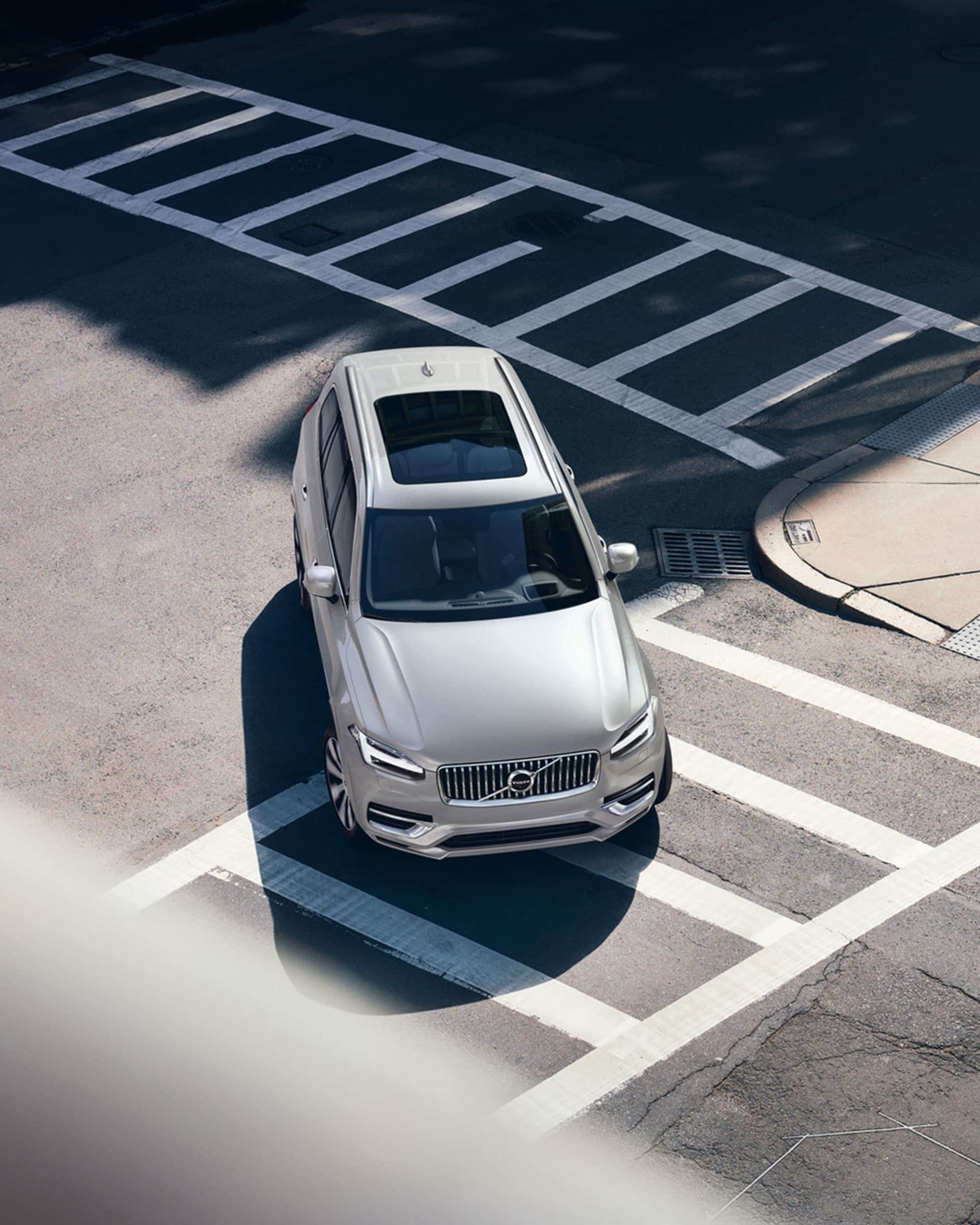 An overhead view shows a light-colored Volvo parked on a city street with white road markings, with shadows from nearby buildings and a fire hydrant visible.