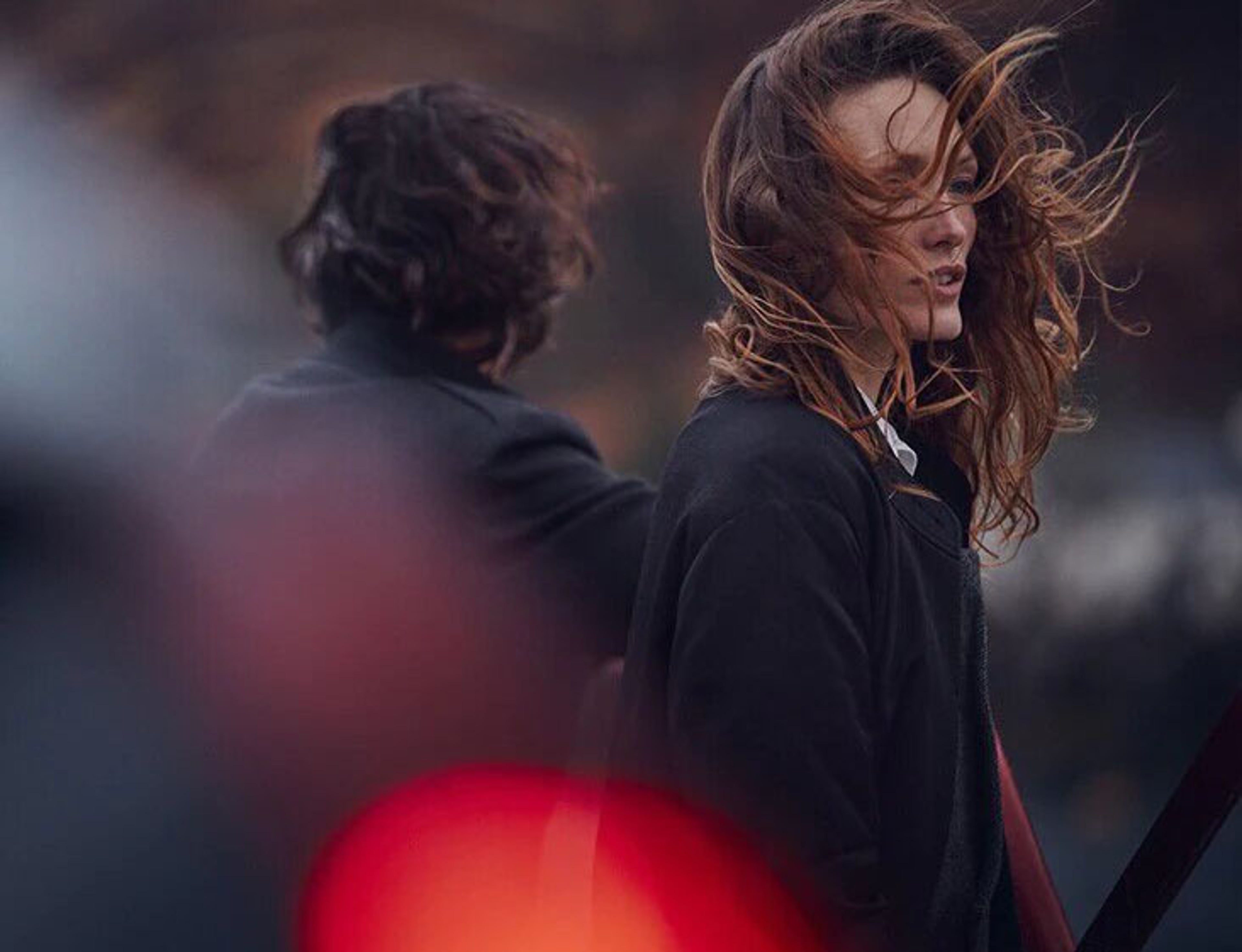 Une femme aux cheveux flottant au vent regarde sur le côté, avec une deuxième personne floue en arrière-plan qui détourne le regard, et un élément rouge flou au premier plan.