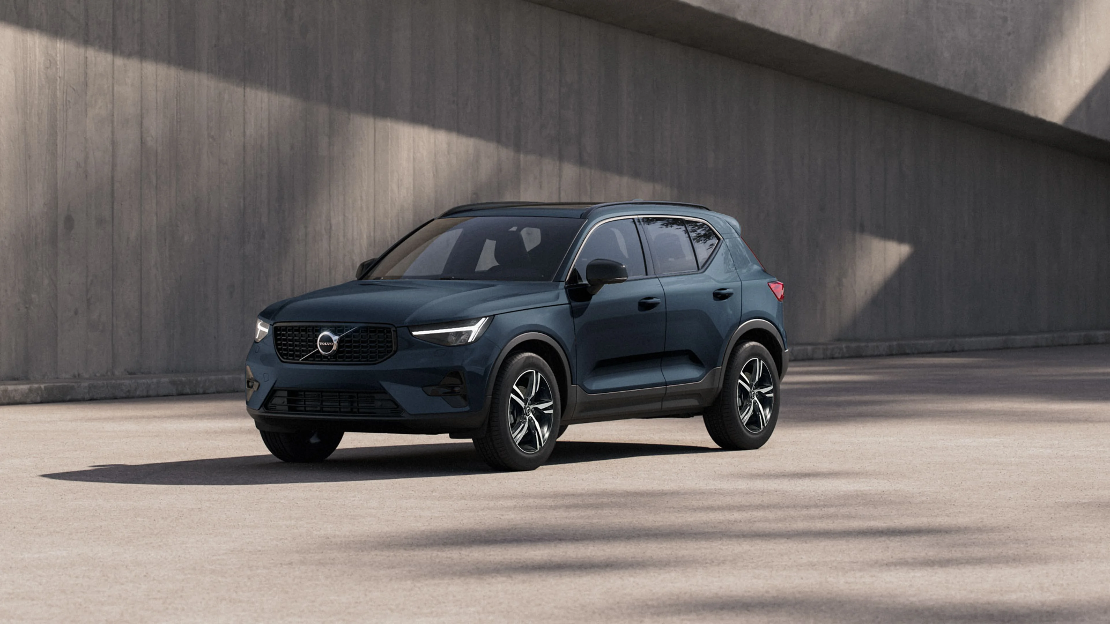 SUV Volvo bleu Denim garé sur une surface en béton, avec un mur gris en arrière-plan. Le véhicule est vu en angle.