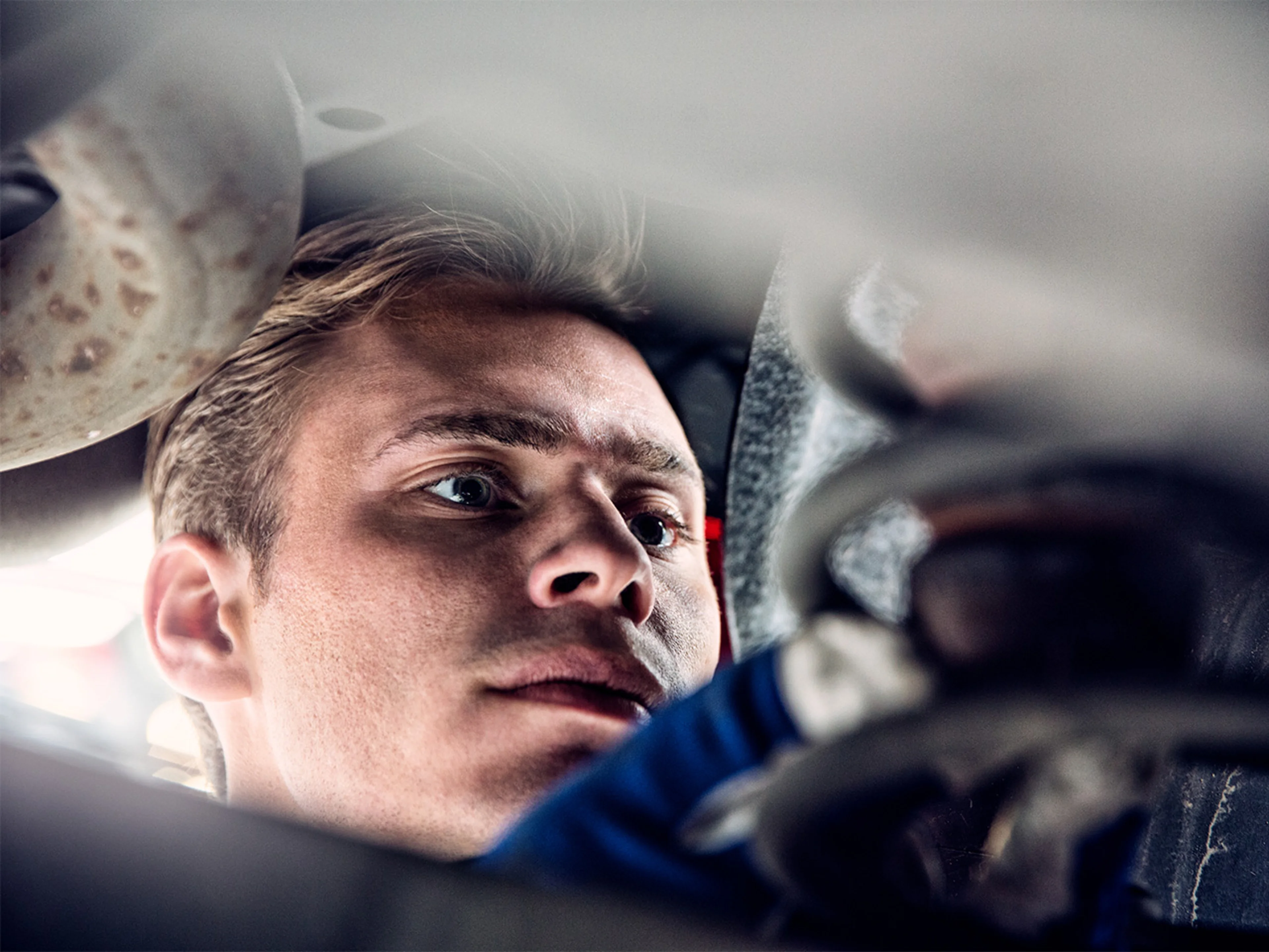 A Volvo technician is working under a vehicle, with mechanical components like a spring visible.