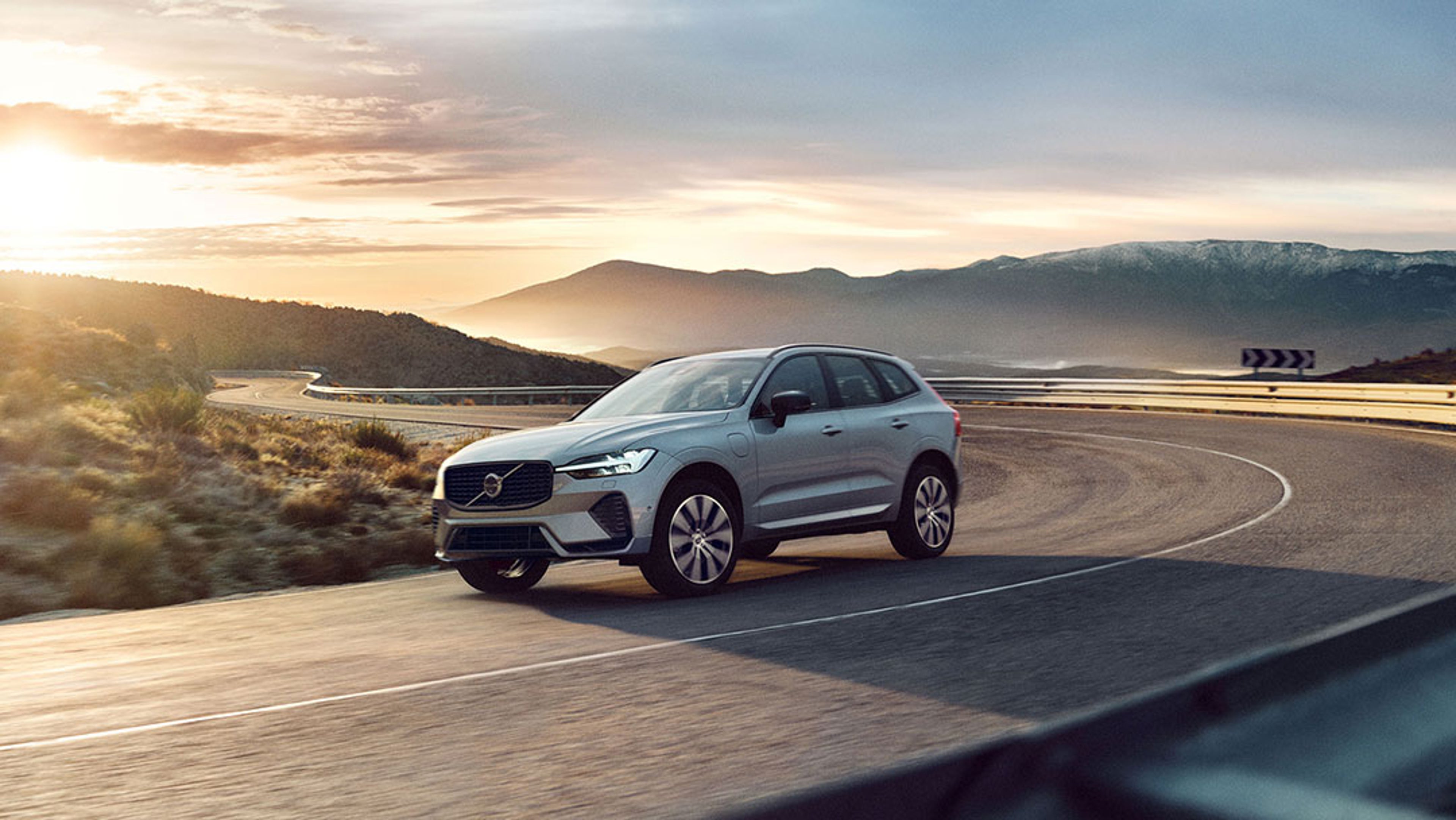 Ein silberner Volvo XC60 fährt auf einer kurvigen, sonnenbeleuchteten Straße durch eine dramatische Landschaft.