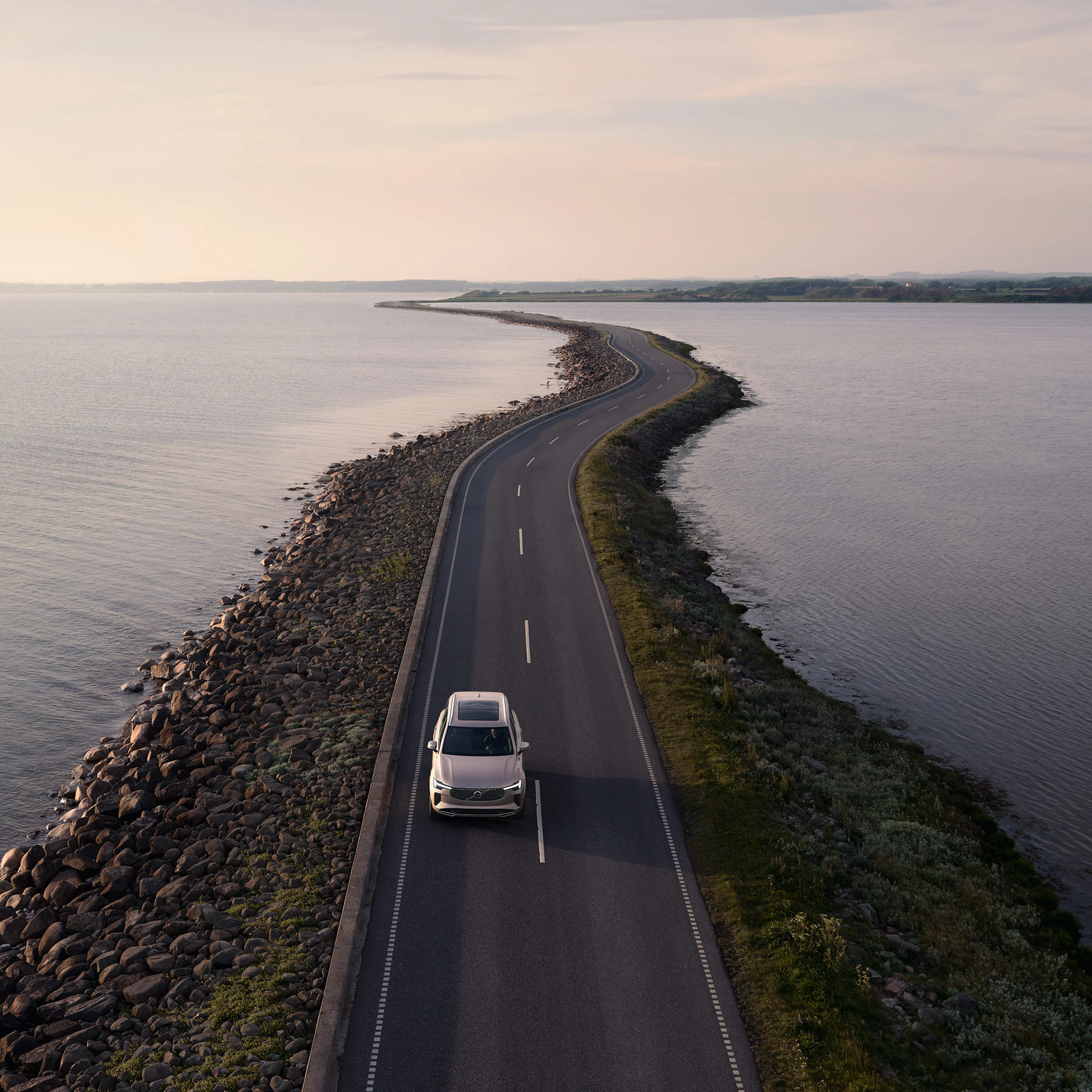 Bright dusk -värinen Volvo XC90 lataushybridikatumaasturi ajaa vesistön pengerrykselle rakennetulla maantiellä.