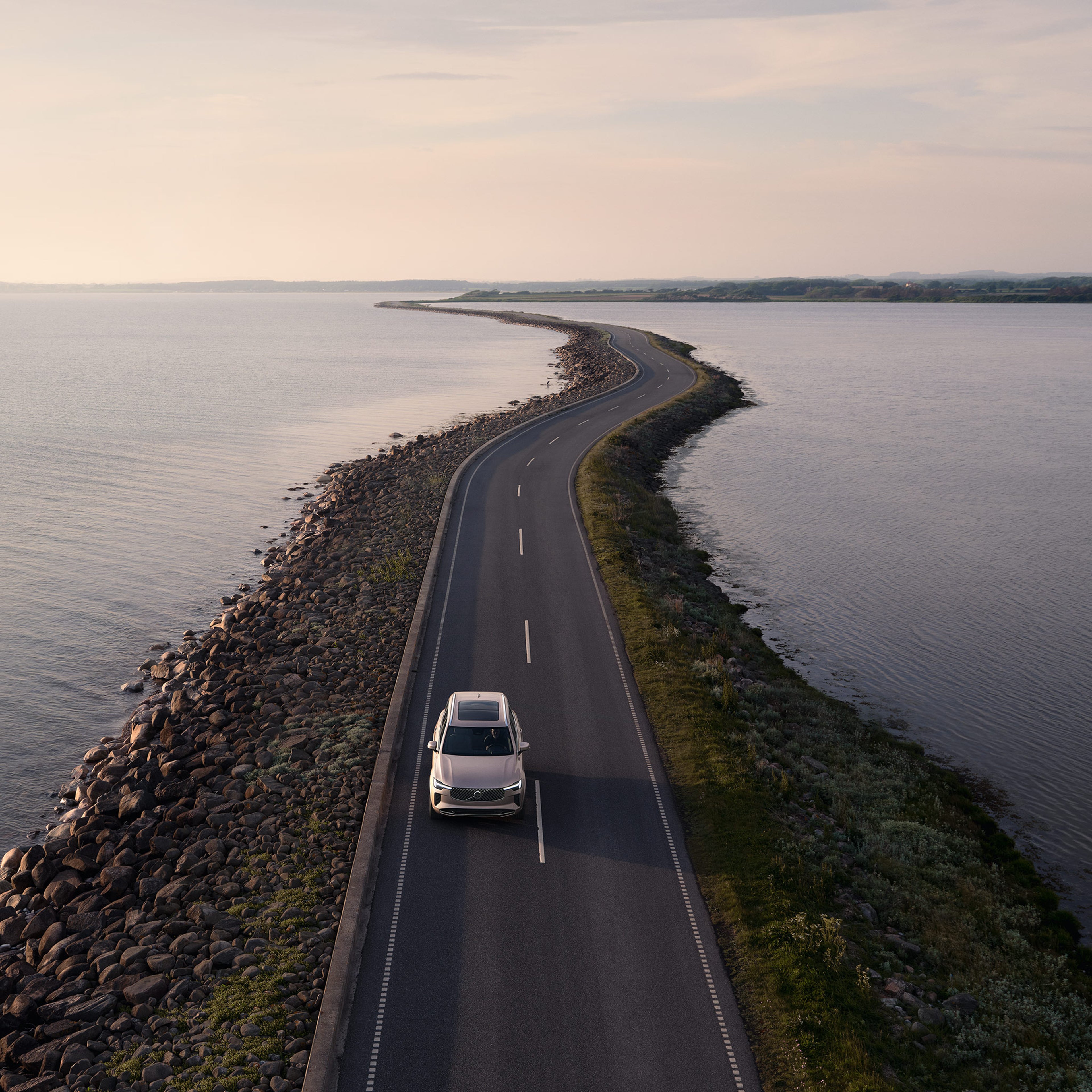 Bright dusk -värinen Volvo XC90 lataushybridikatumaasturi ajaa vesistön pengerrykselle rakennetulla maantiellä.