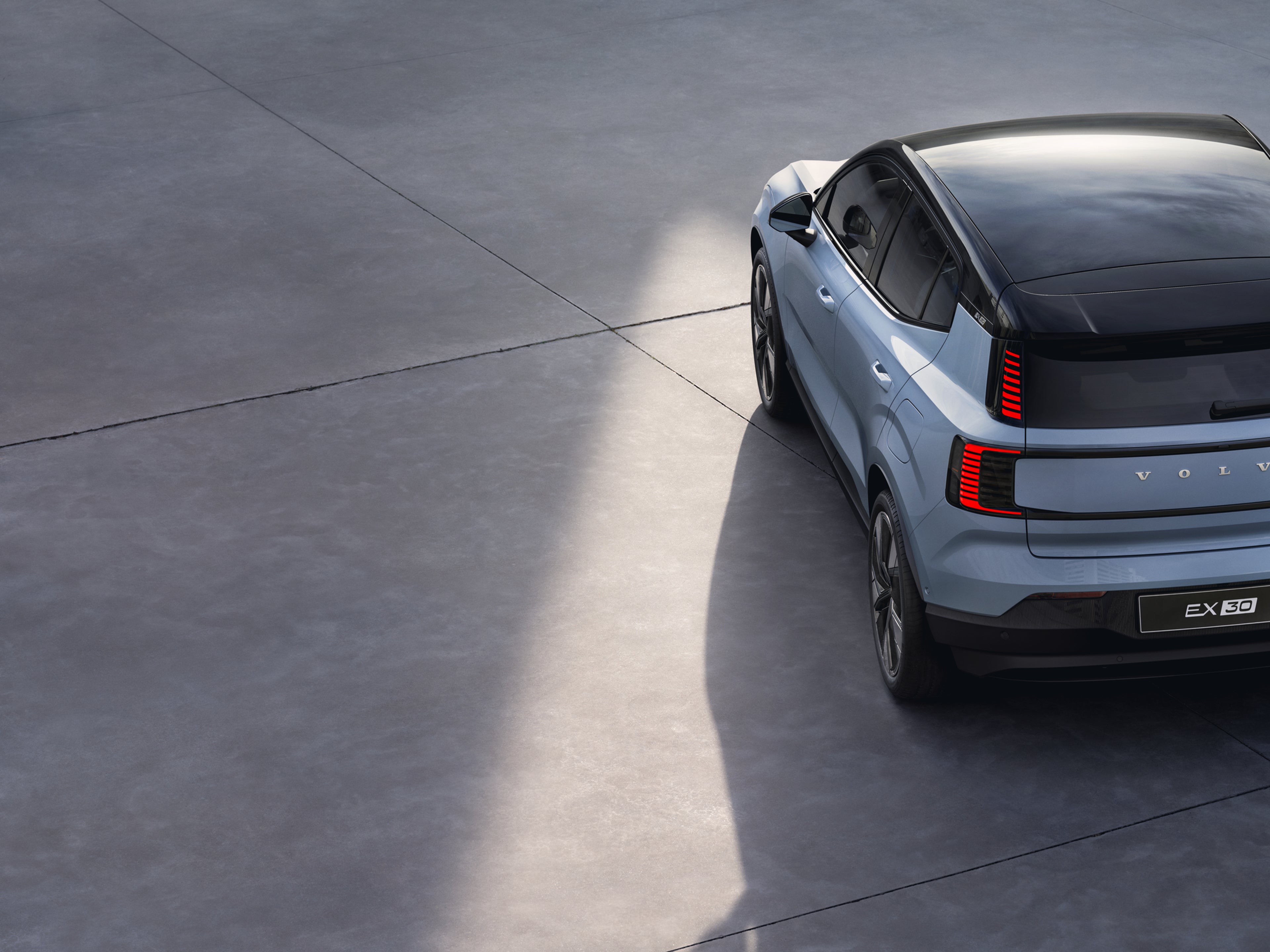A cloud blue, fully electric Volvo EX30 SUV is parked on a smooth concrete surface, pictured from above and behind in sunlight.