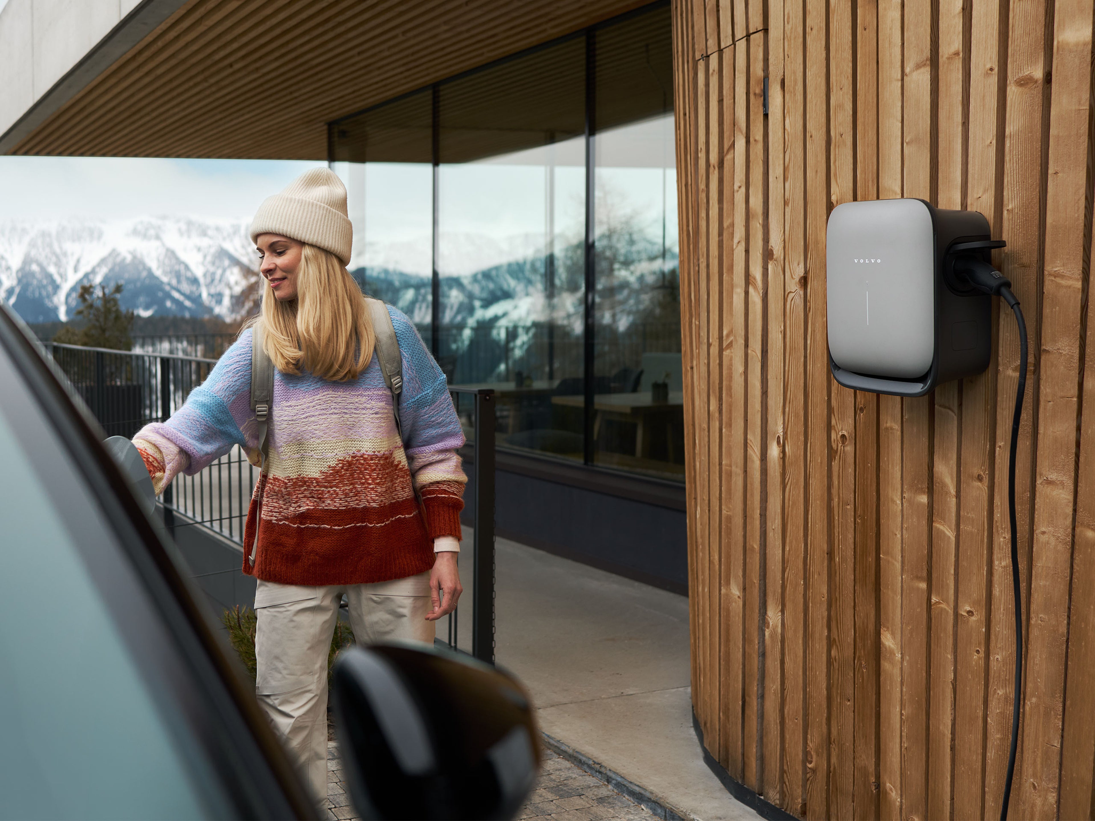 Volvon latausasema kiinnitettynä vuoristohuvilan laudoitettuun seinään. Pipoon ja villapaitaan pukeutunut nainen asettaa latauspistoketta täyssähköisen Volvon latausrasiaan.
