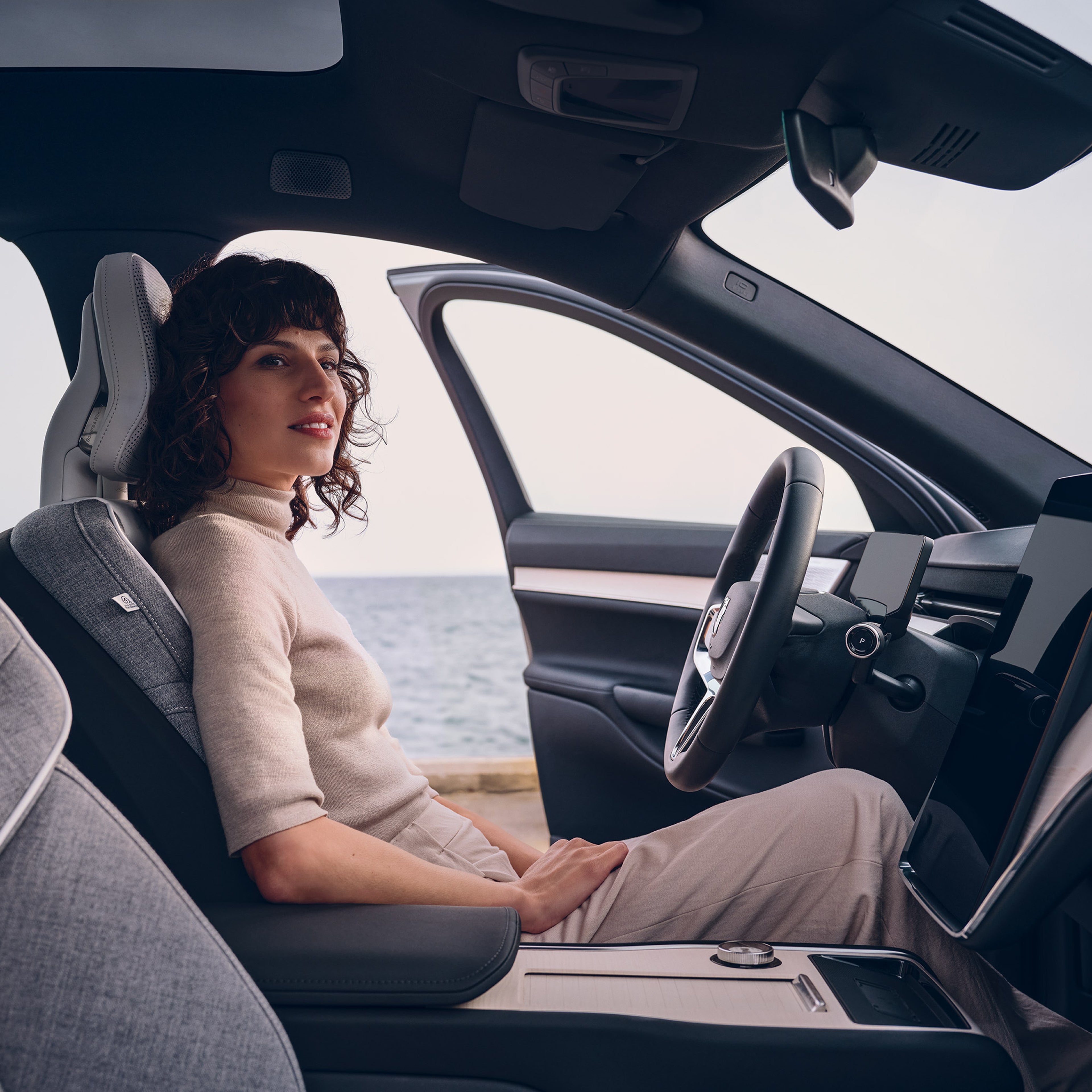 Nainen istuu Volvo EX90 täyssähköisen katumaasturin kuskin paikalla ja auton etuovi on auki, taustalla näkyy meri.