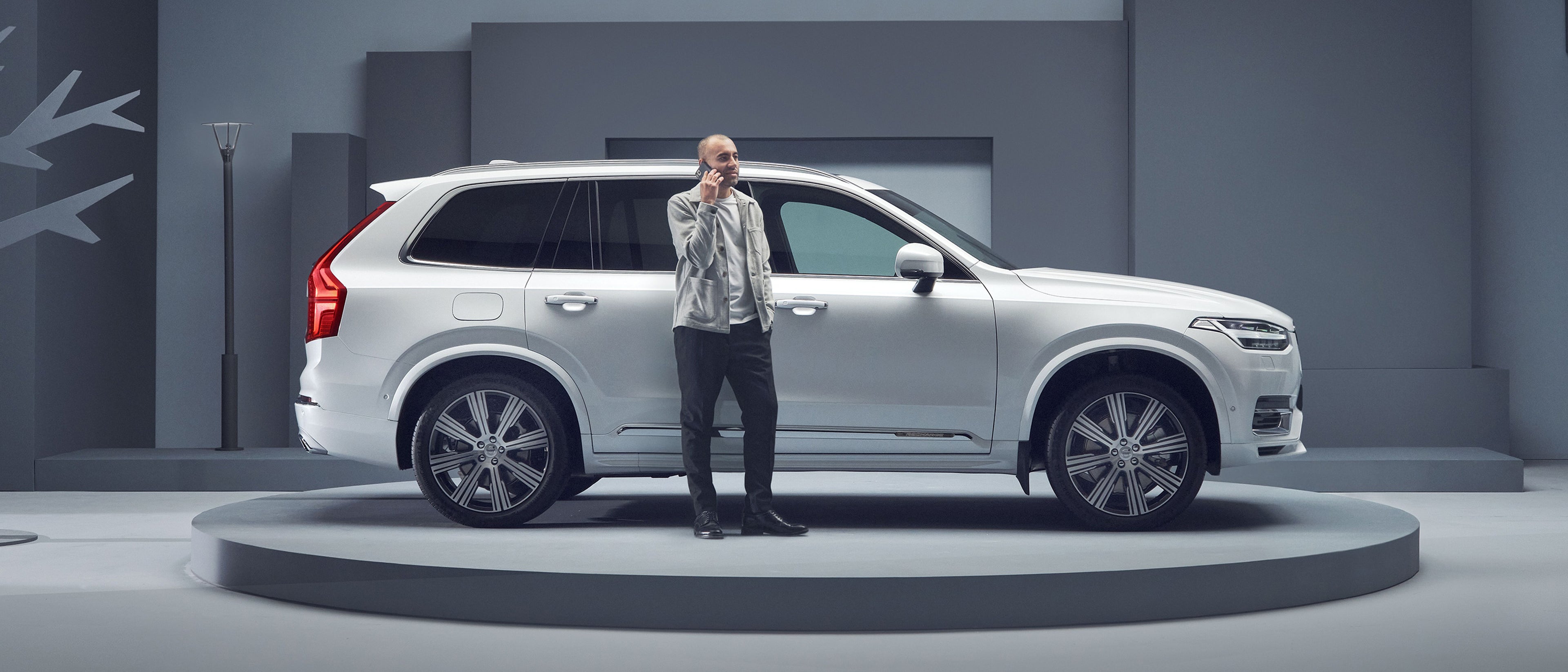 A white Volvo XC90 plug in hybrid SUV displayed on a circular platform against a minimalist backdrop, with customer who is on his phone.