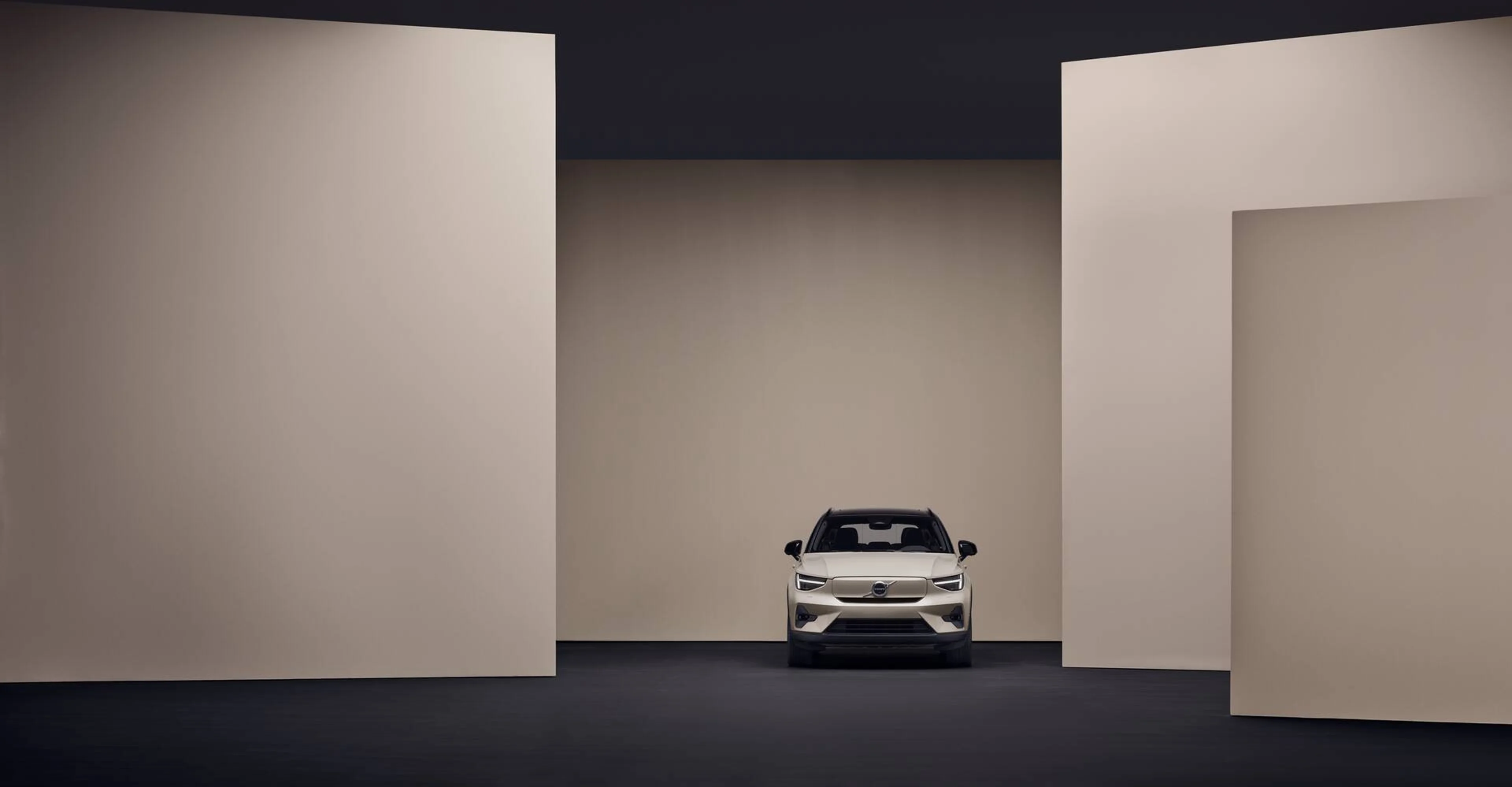 En elegant, hvid EX40 Volvo SUV er centralt placeret i et minimalistisk, geometrisk rum med beige vægge og mørkt gulv, hvilket giver en moderne og sofistikeret stemning.