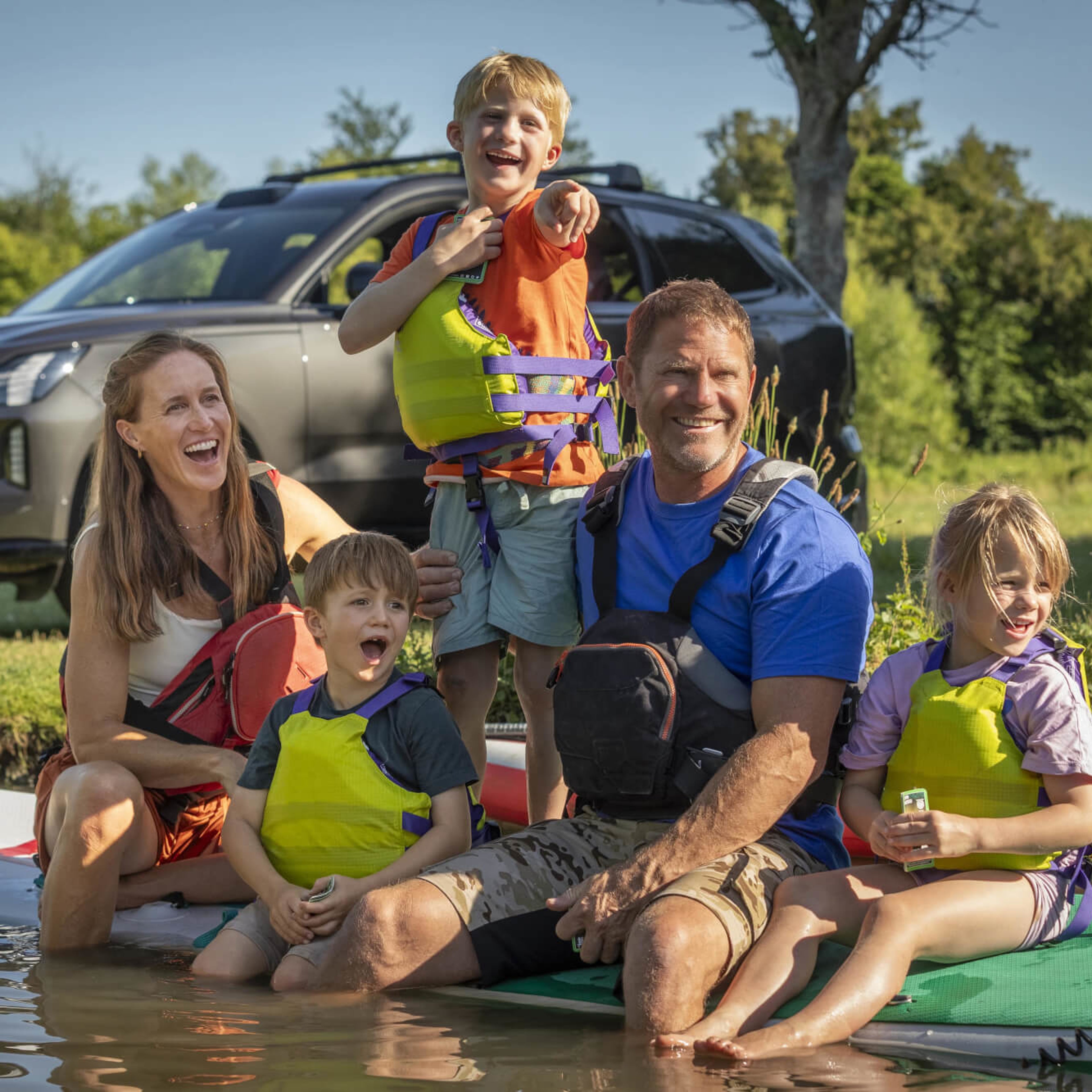 Steve Backshall with his family laughing on a paddleboard with Volvo EX90 parked in the background