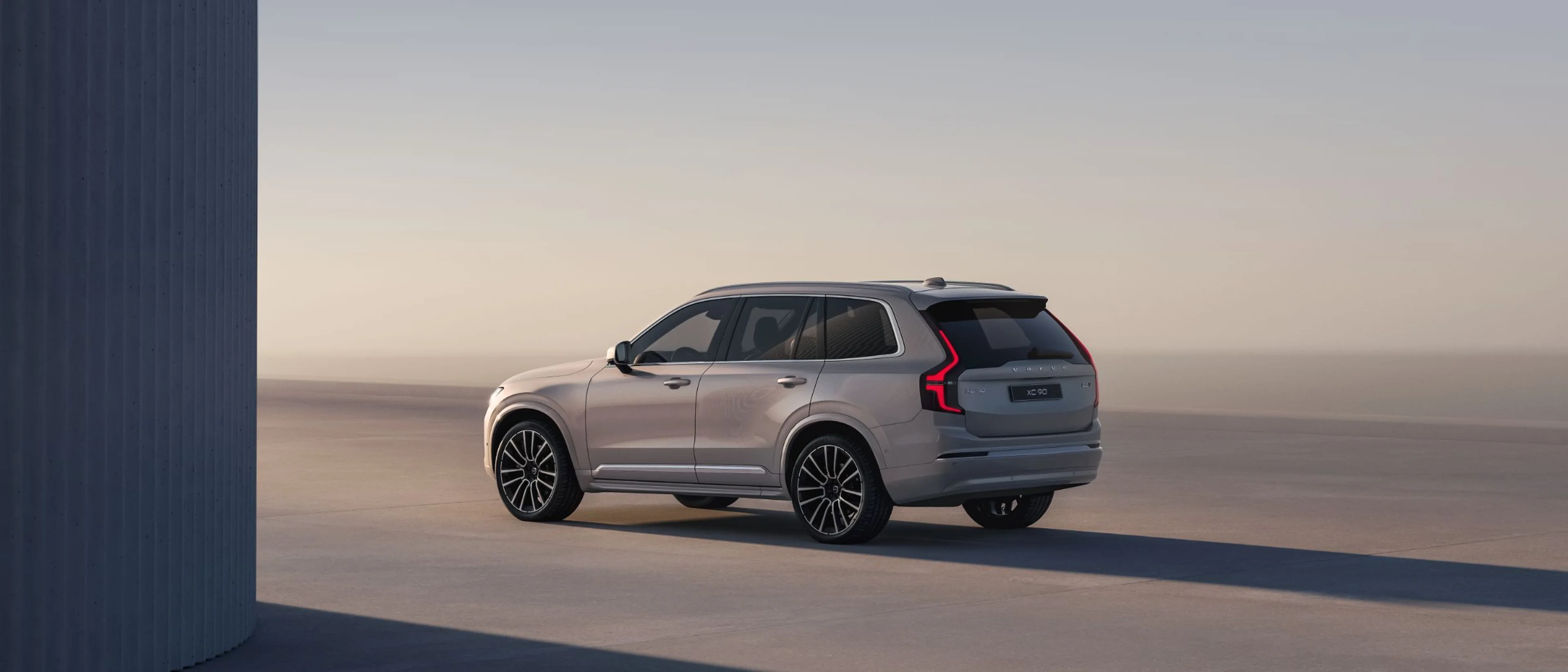 Сребрен Volvo XC90 SUV паркиран на отворен простор, заден три-четвртински поглед.