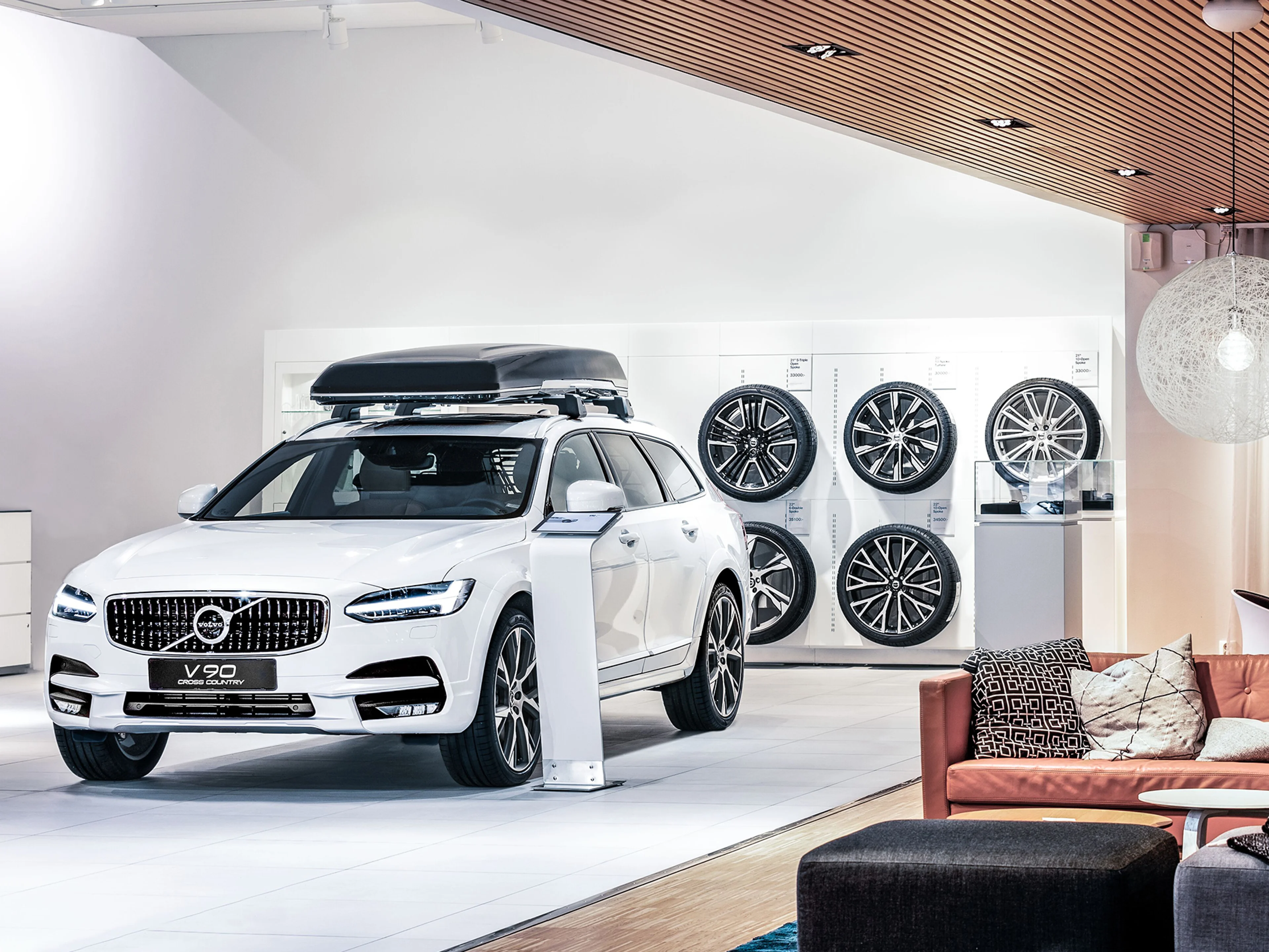 Ein weißer Volvo V90 mit Dachbox steht in einem Showroom vor einer Wand mit Volvo Felgen.