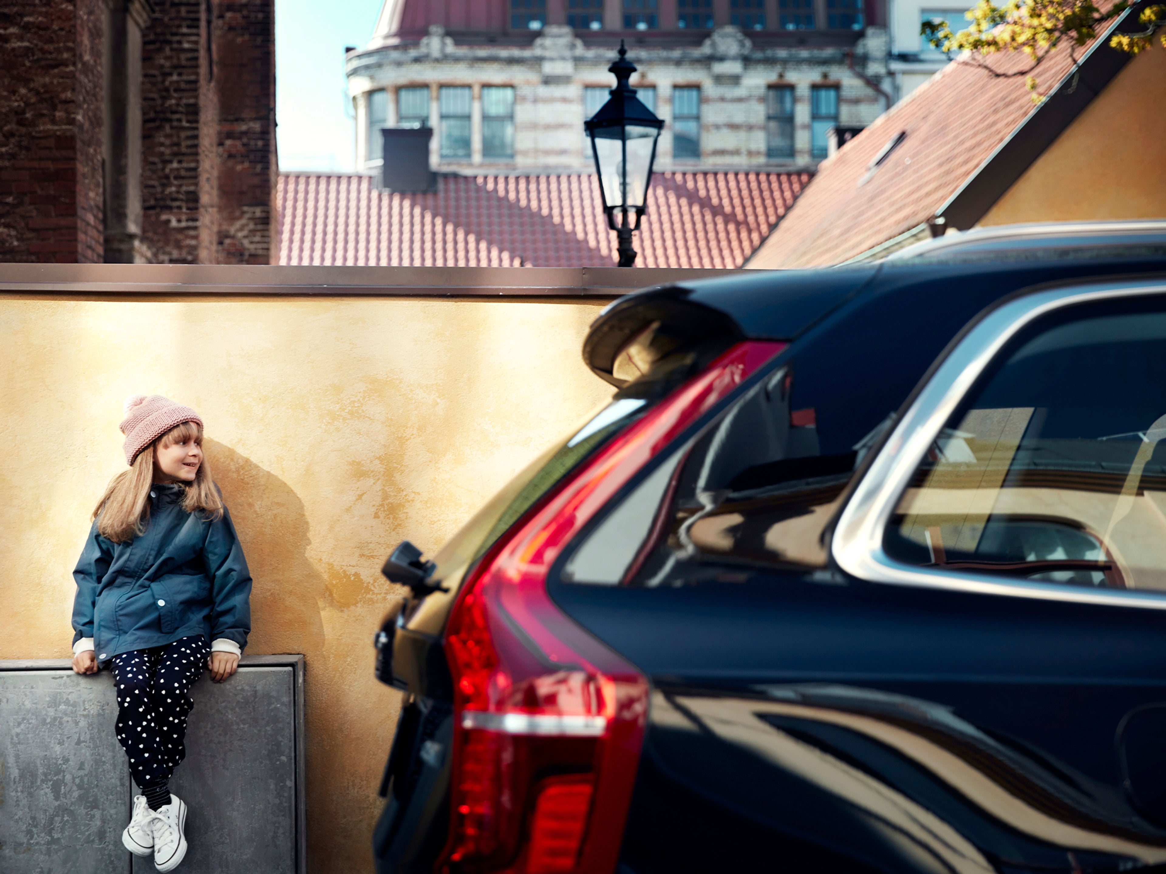 Una niña recargada en la pared con un vehículo Volvo XC90 estacionado al lado.