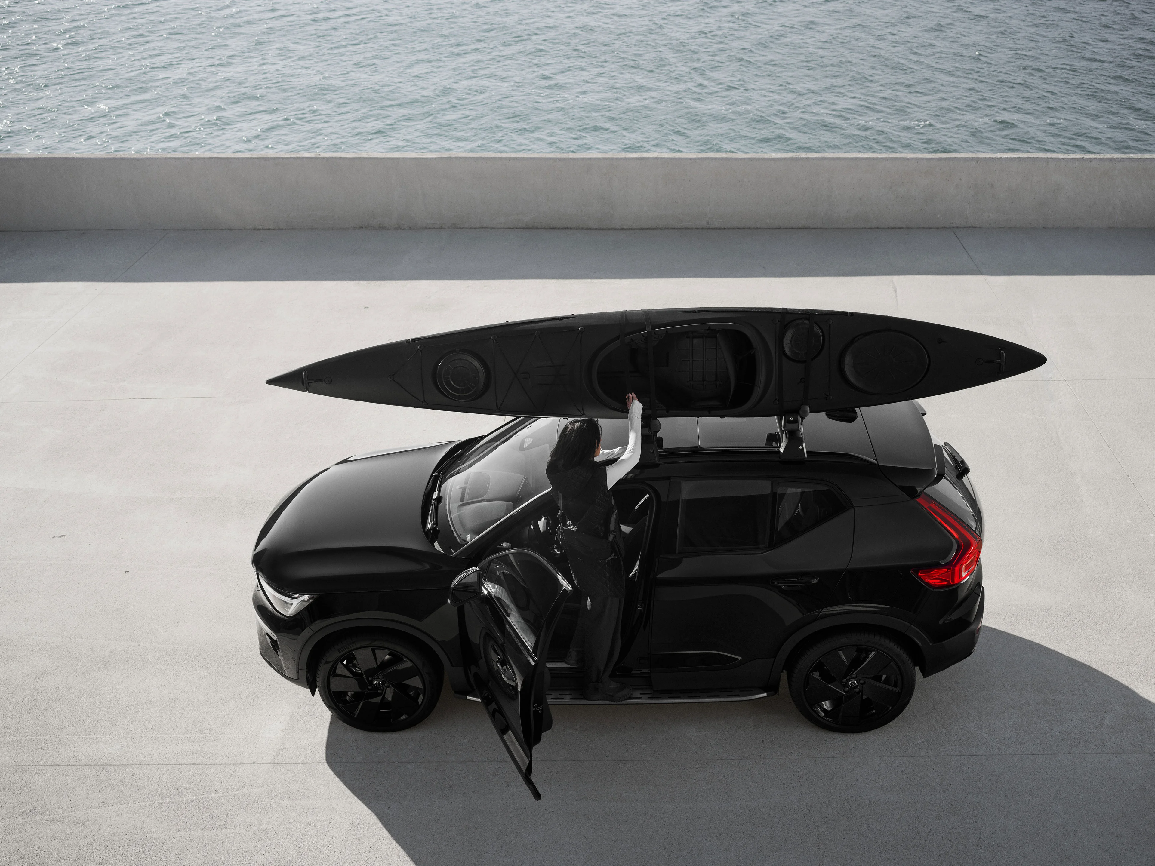 Eine Frau montiert ein schwarzes Kajak auf dem Dachträger eines Volvo XC40 Black Edition, der parallel zu einem Gewässer auf einer Brücke parkt.