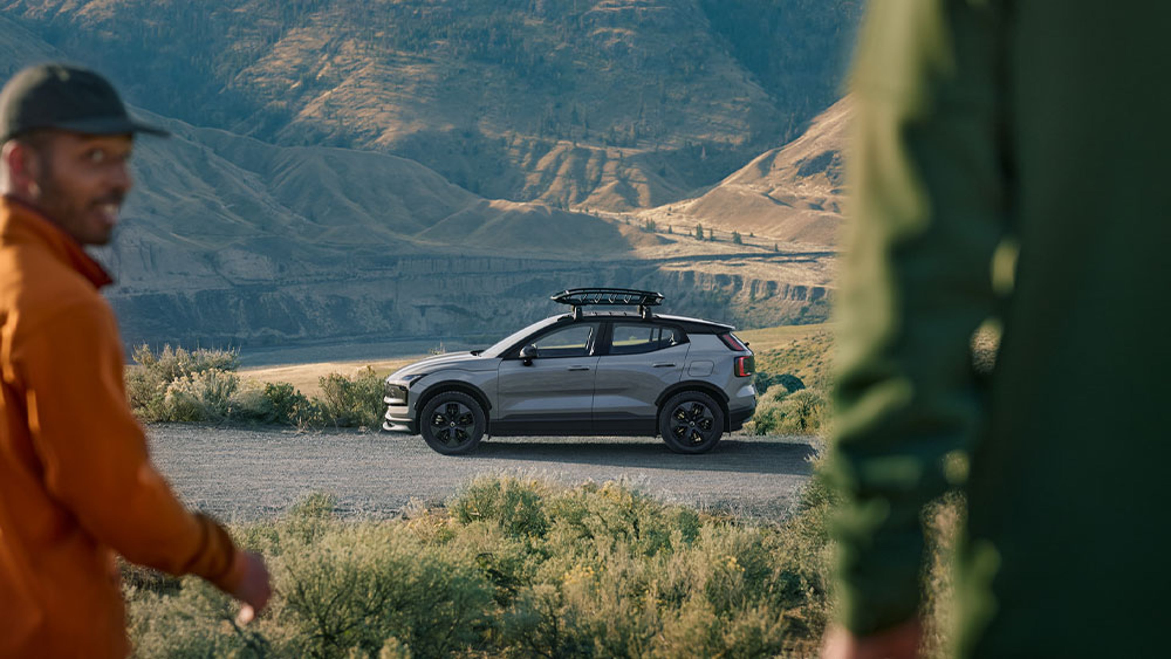 Volvo EX30 gris garé sur une route en gravier avec montagnes en arrière-plan et deux silhouettes floues au premier plan