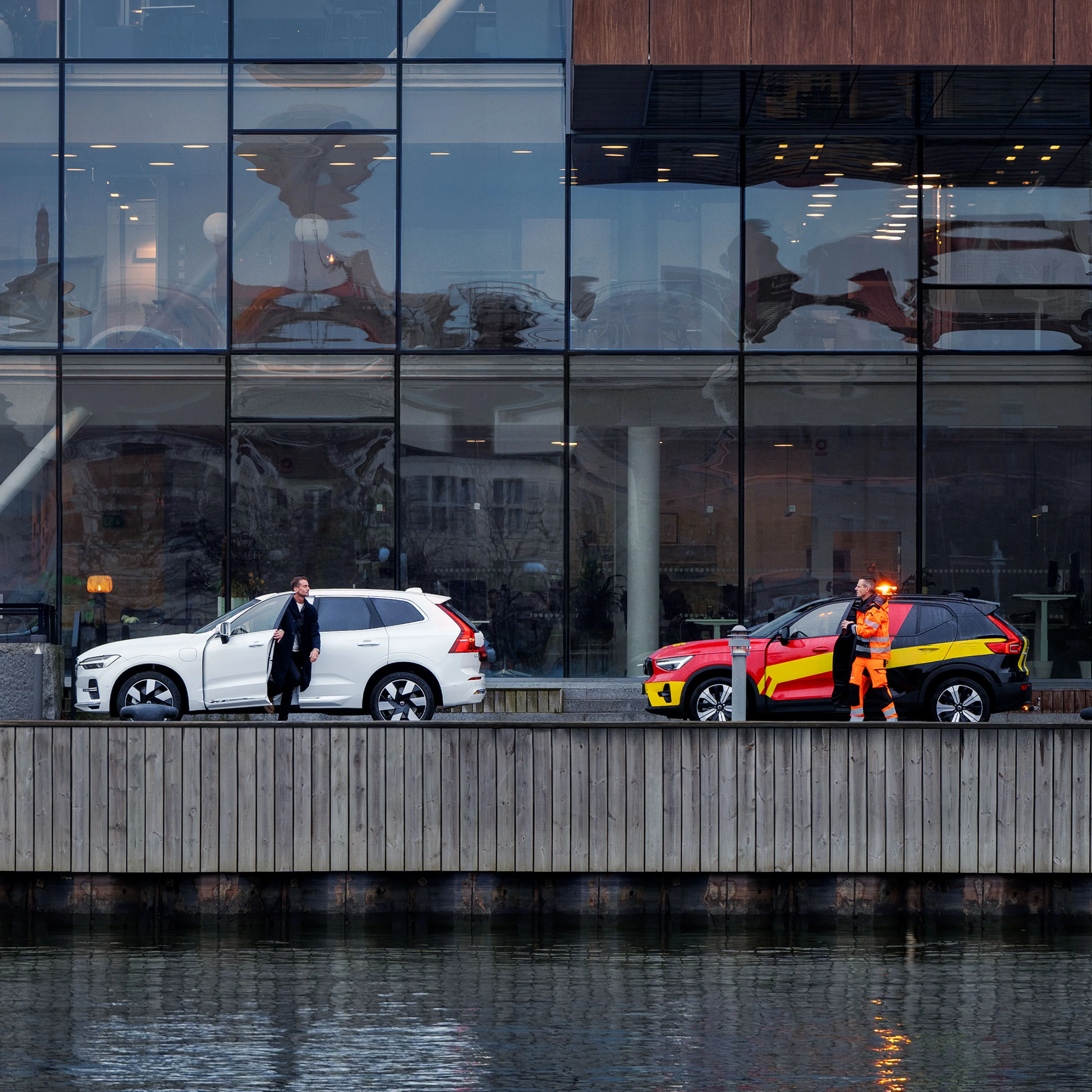 Valkoinen Volvo ja tiepalvelun auto pysäköitynä veden äärellä sijaitsevan toimistotalon edustalle.