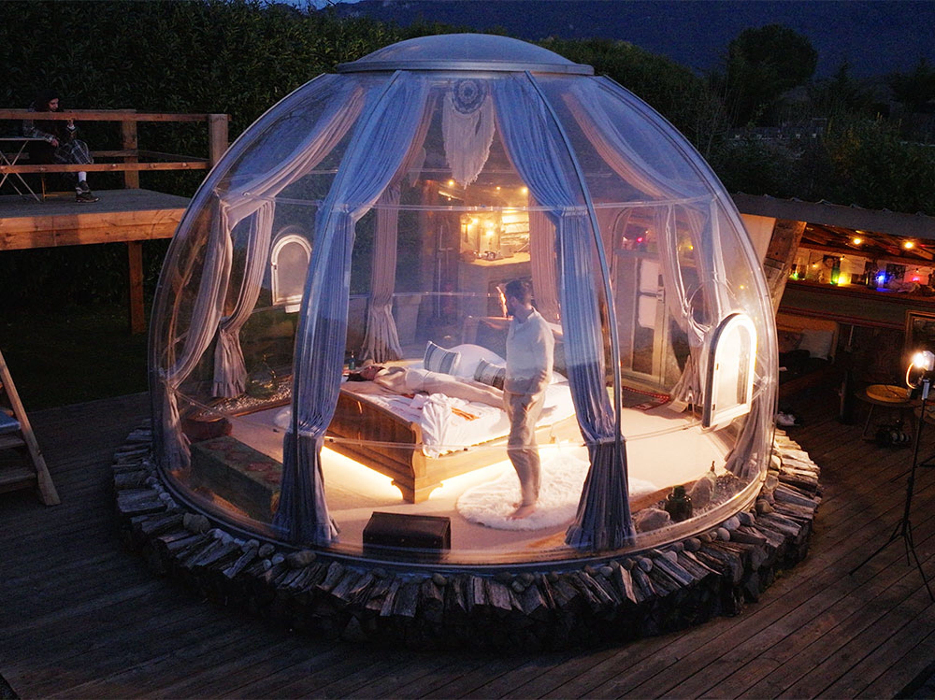Chambre en dôme transparent avec lit et personne, entourée d’une terrasse en bois et d’un coin salon extérieur