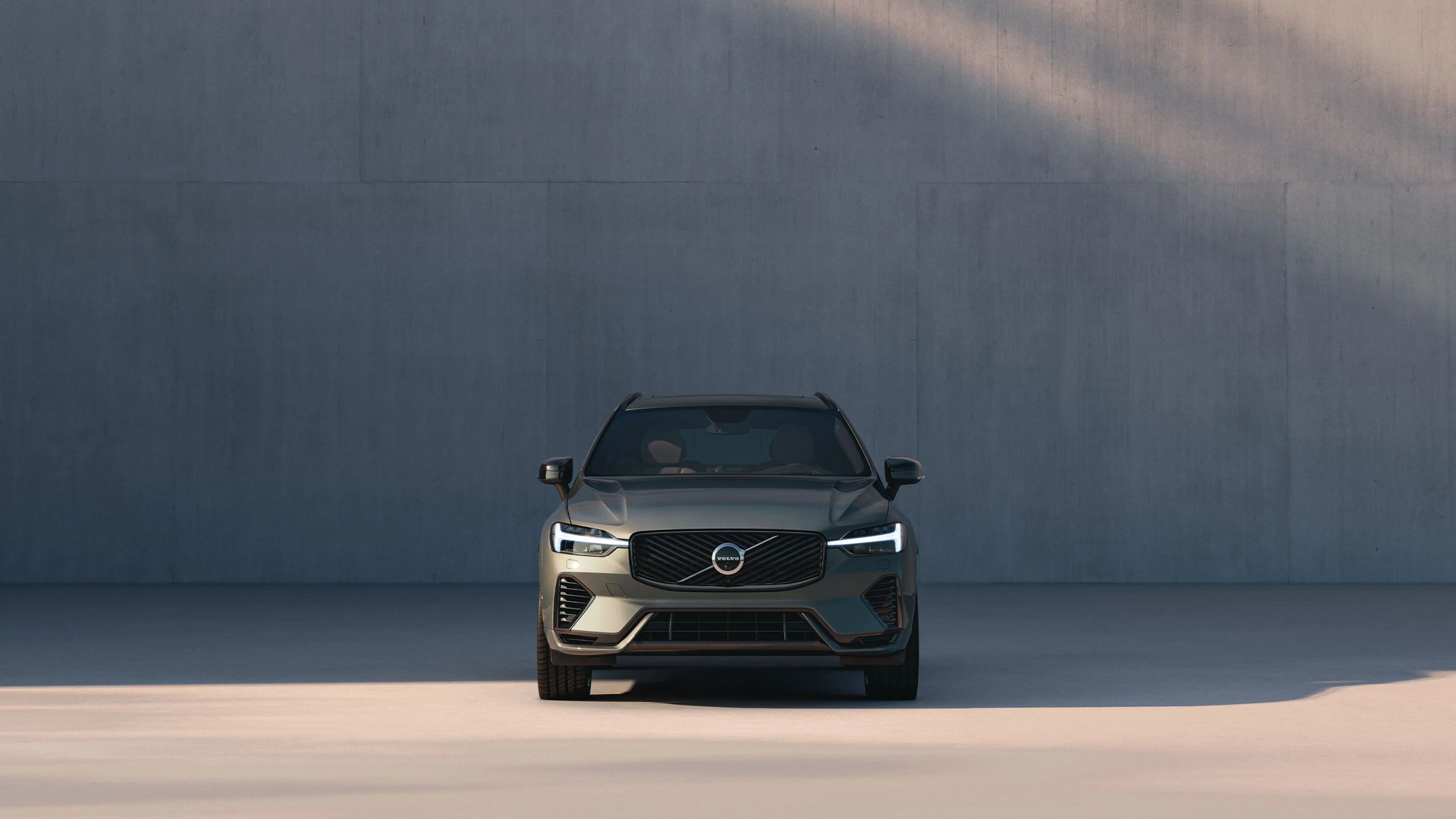 Ein graphitfarbener Volvo XC60, der von links von der Sonne beleuchtet wird, parkt vor einer Betonwand.