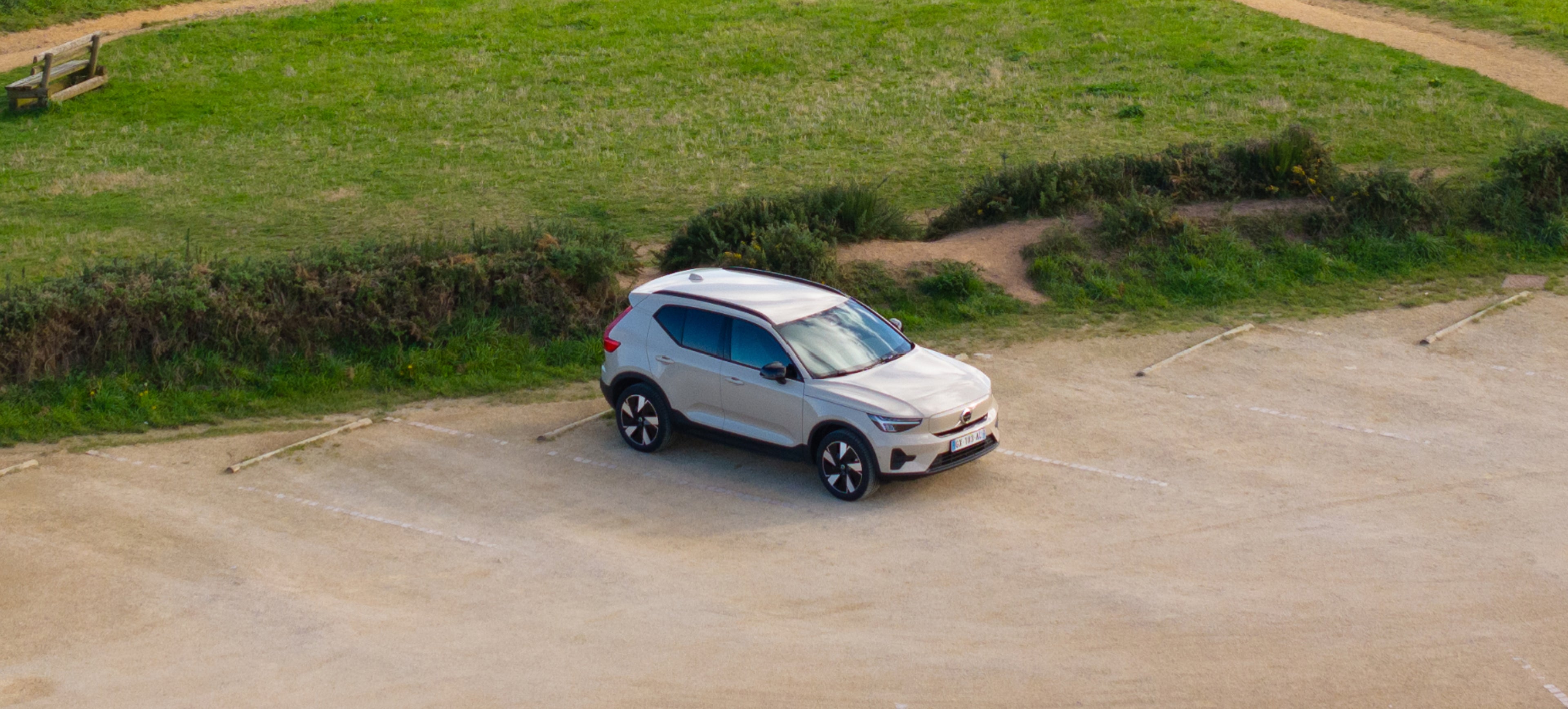 Voiture argentée sur parking en terre près d’un sentier herbeux, vue sur l’océan et rochers au loin.
