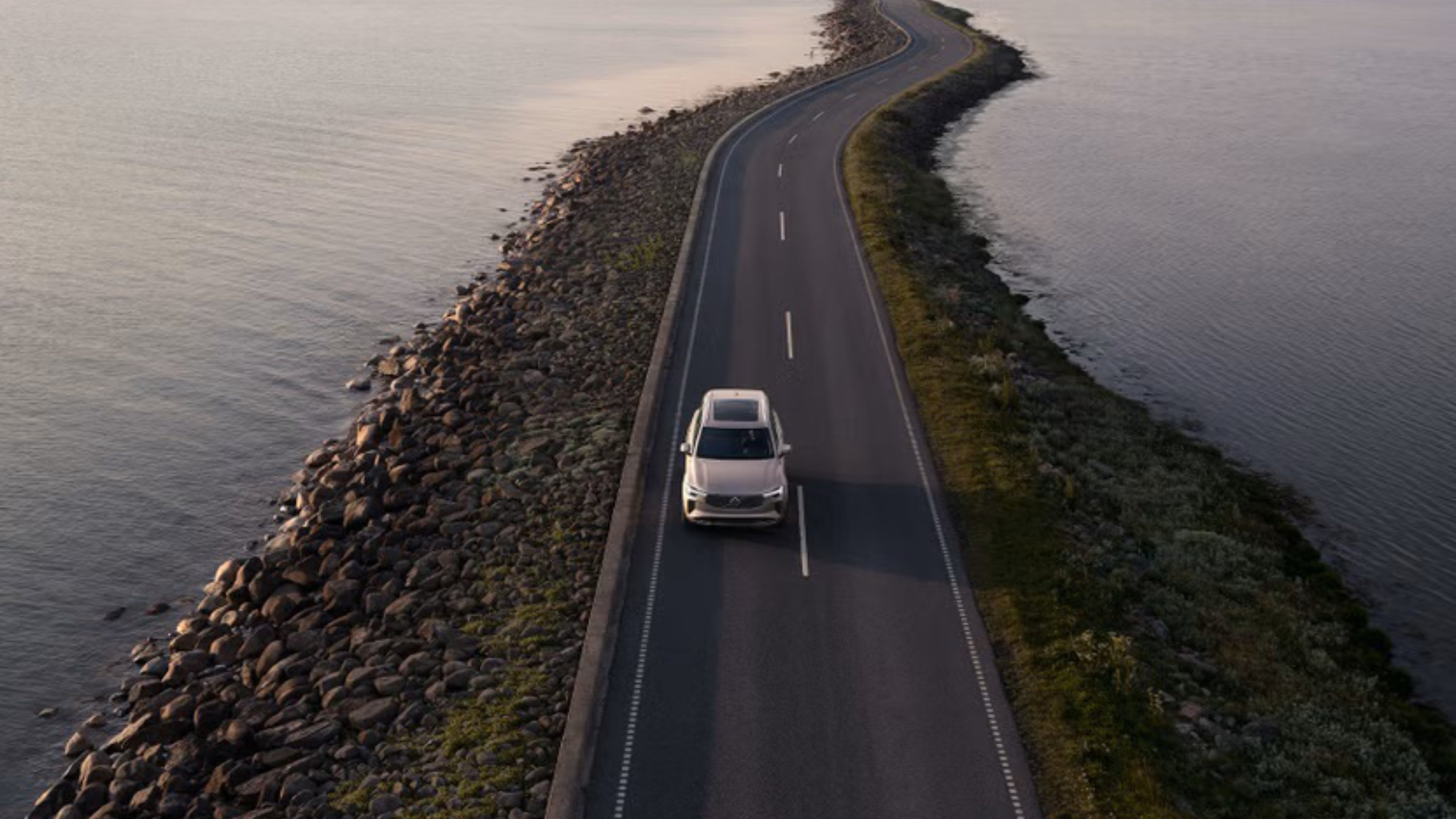 Frontalansicht aus der Luft eines silbernen Volvo V90 der auf einer Straße umgeben von Wasser fährt.