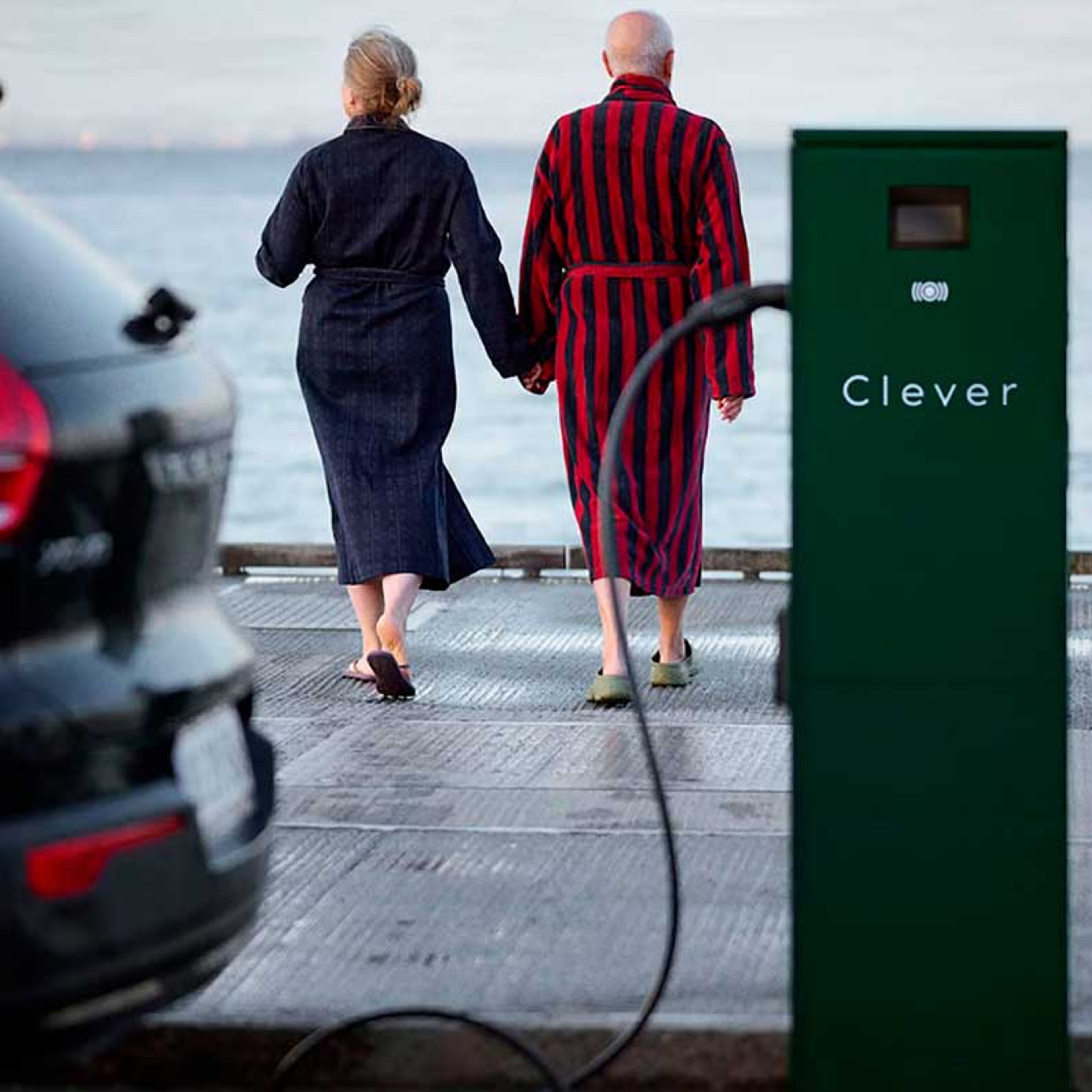 Et par i badekåber går hånd i hånd mod havet. En Volvo elbil lader op ved en nærliggende ladestation med "Clever" skrevet på, hvilket fremkalder en følelse af ro og modernitet.