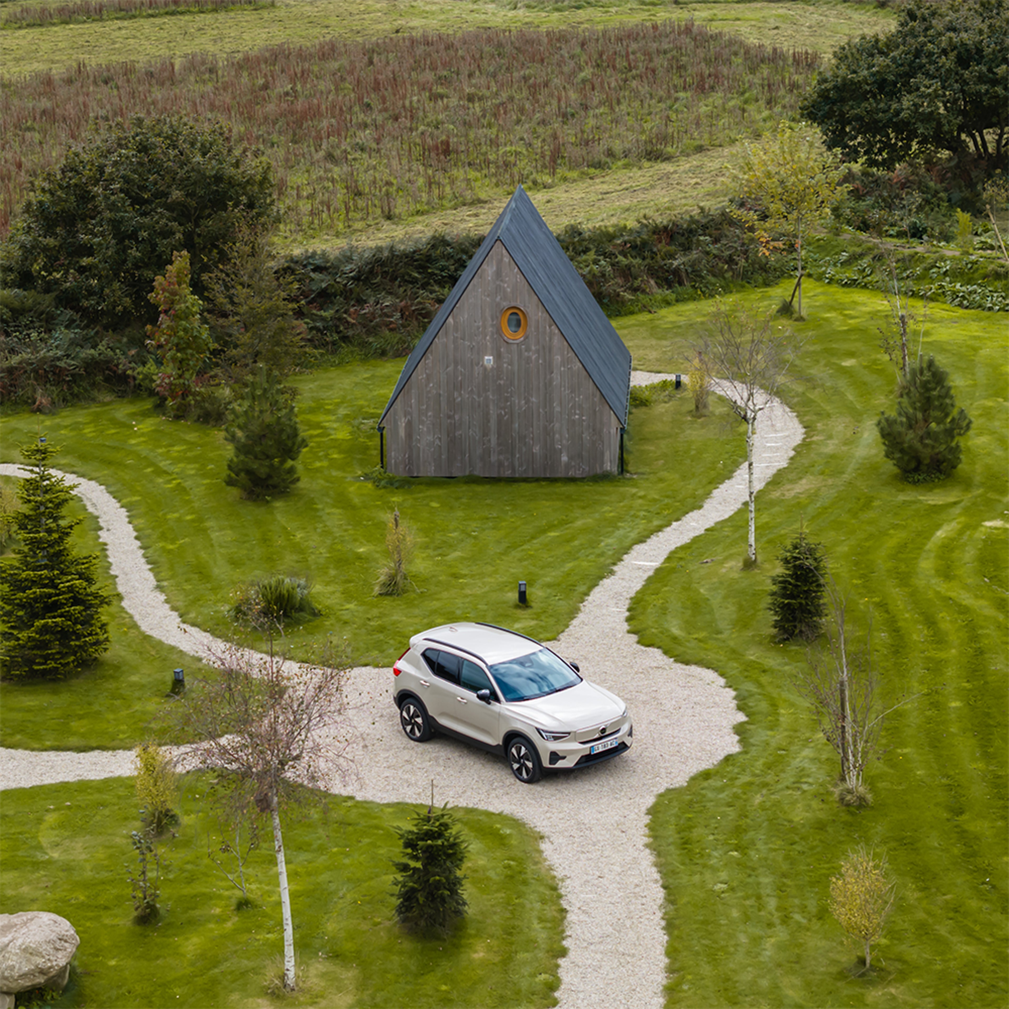 Volvo blanche sur allée en gravier sinueuse, pelouse verte, arbres et cabane en bois en arrière-plan.