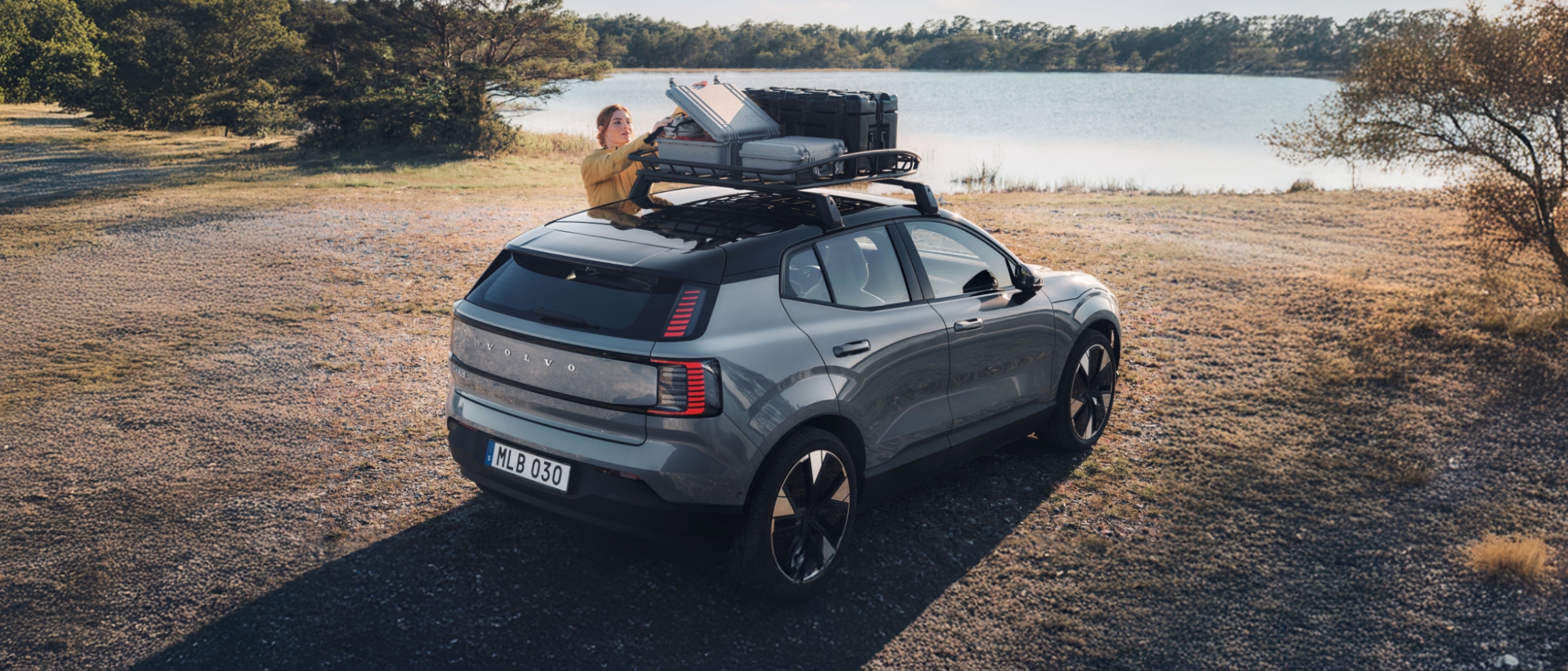 Volvo EX30 parkiran ob jezeru, ženska v rumeni jakni nalaga prtljago na strešni prtljažnik vozila v naravnem okolju.