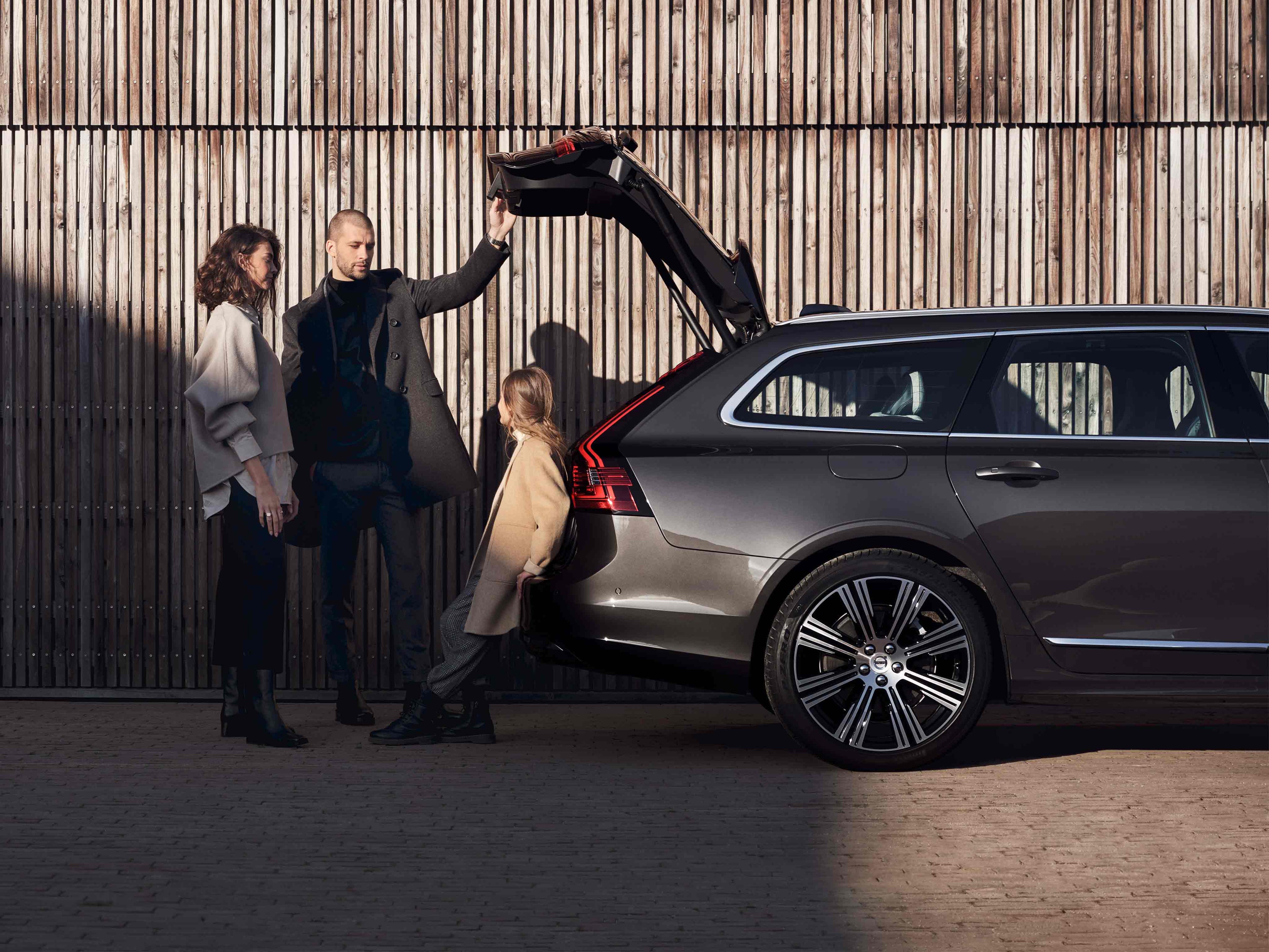 Nainen mies ja lapsi seisoo tummanharmaan Volvo V90:n avonaisen takaluukun alla, taustalla puusäleaita.