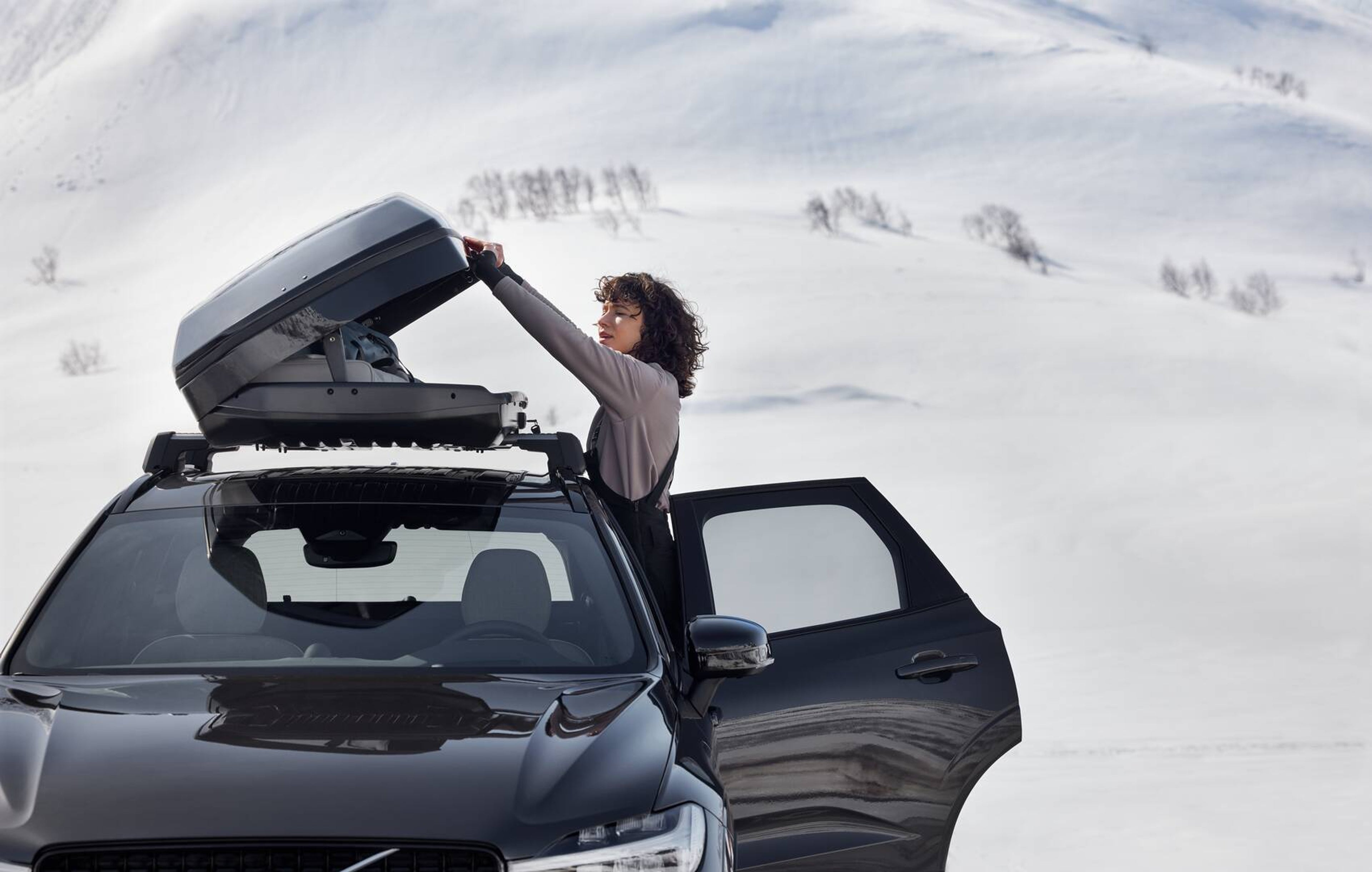 Person with brown hair opening roof box on top of their Volvo car in front of a snowy mountain
