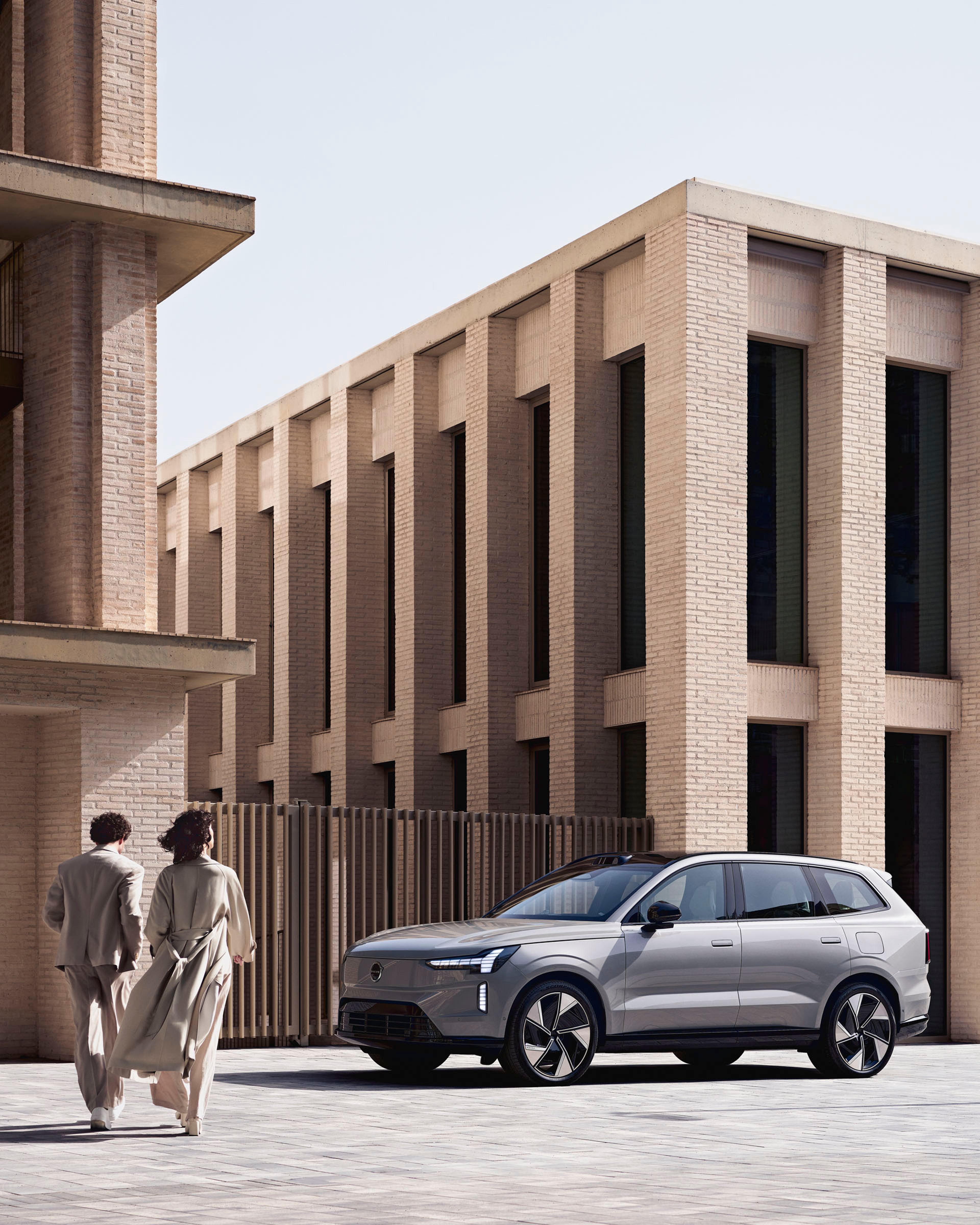 Dos personas caminando hacia un Volvo EX90 eléctrico aparcado entre edificios.
