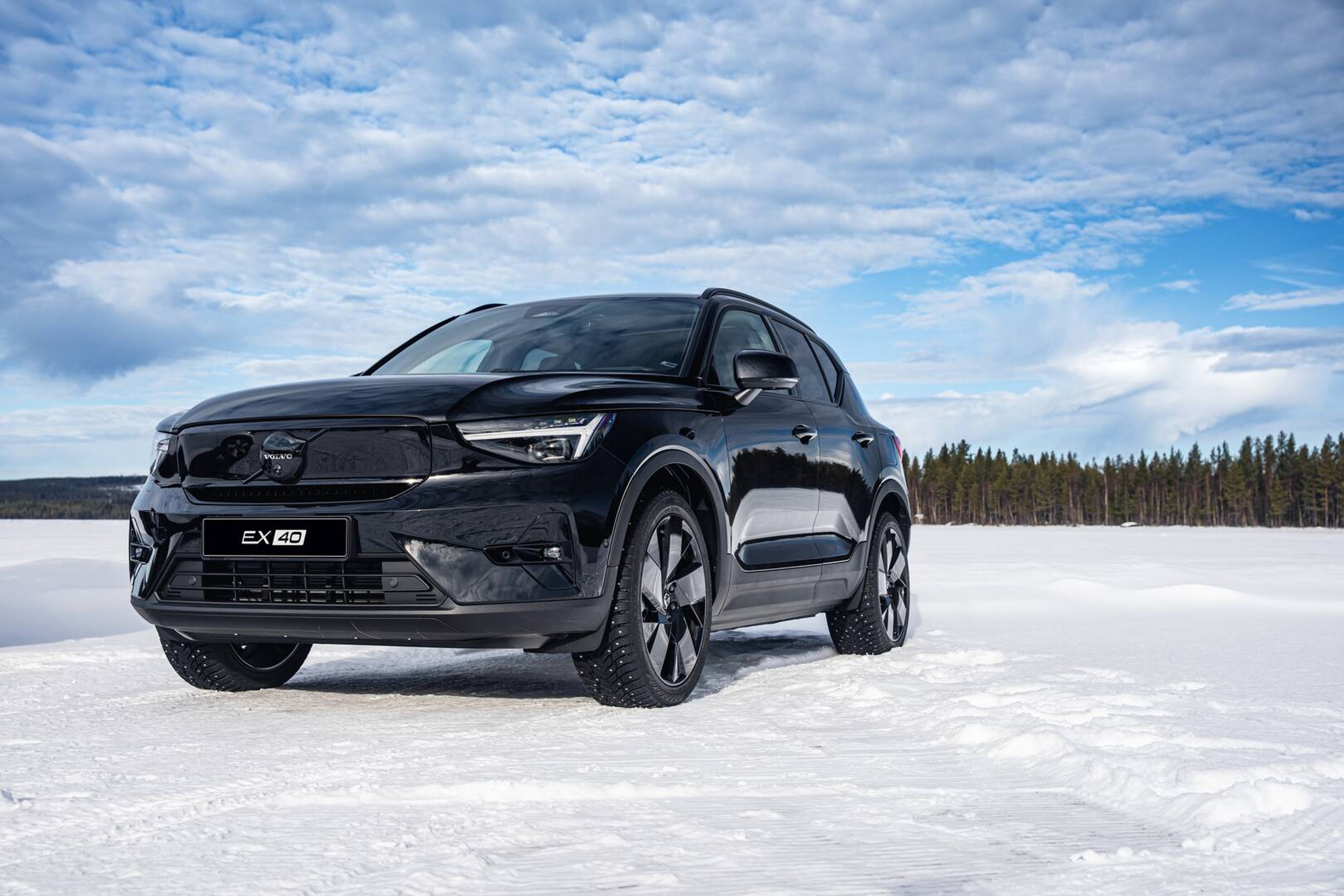 Sort Volvo SUV med mærket "EX40" parkeret i sneen under en delvist skyet himmel. Omgivende træer og det rolige landskab fremkalder en følelse af eventyr.