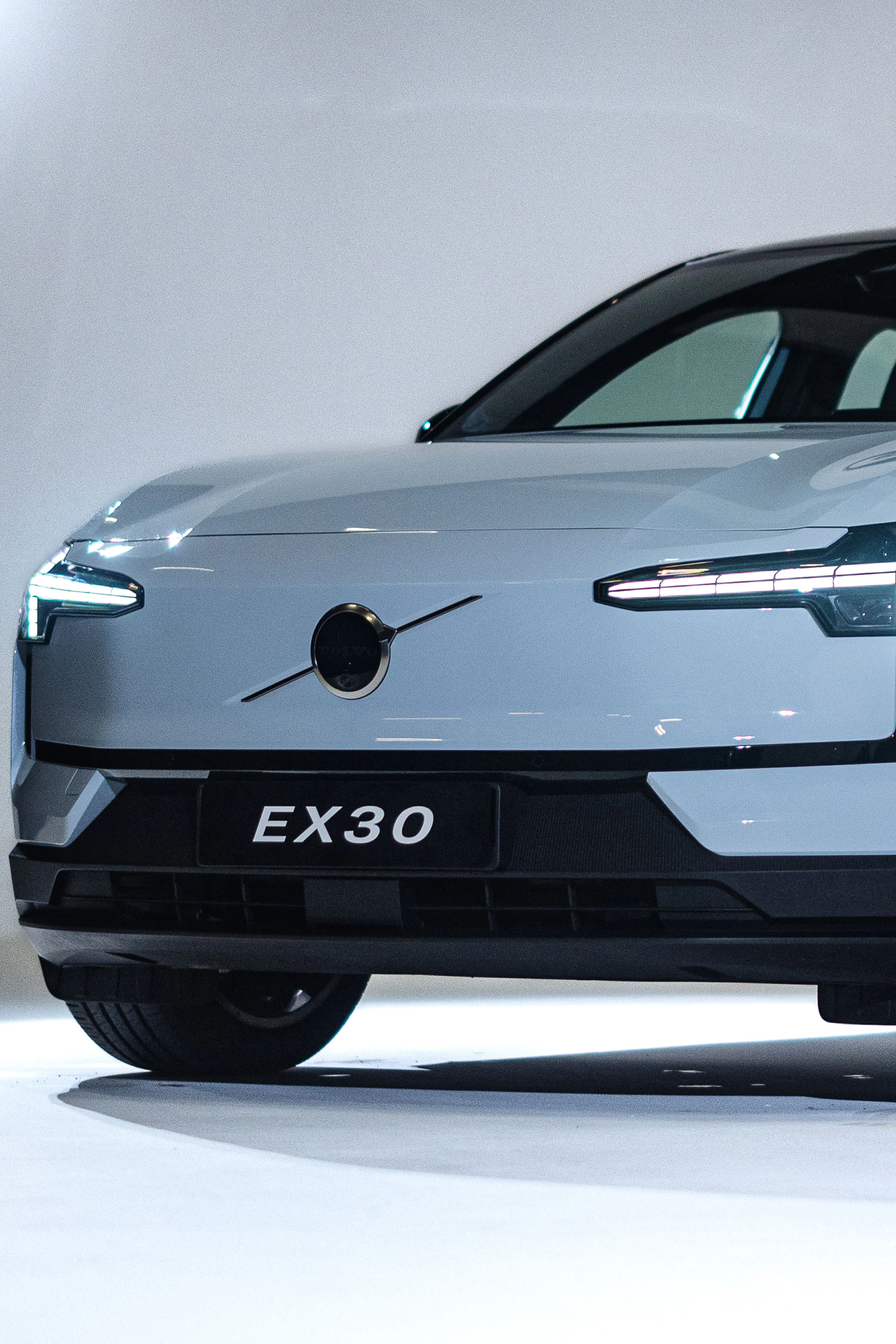 Fronten af en elegant sølvfarvet Volvo EX30, der fremhæver moderne LED-forlygter og et cirkulært emblem, med "EX30" tydeligt vist på nummerpladen.