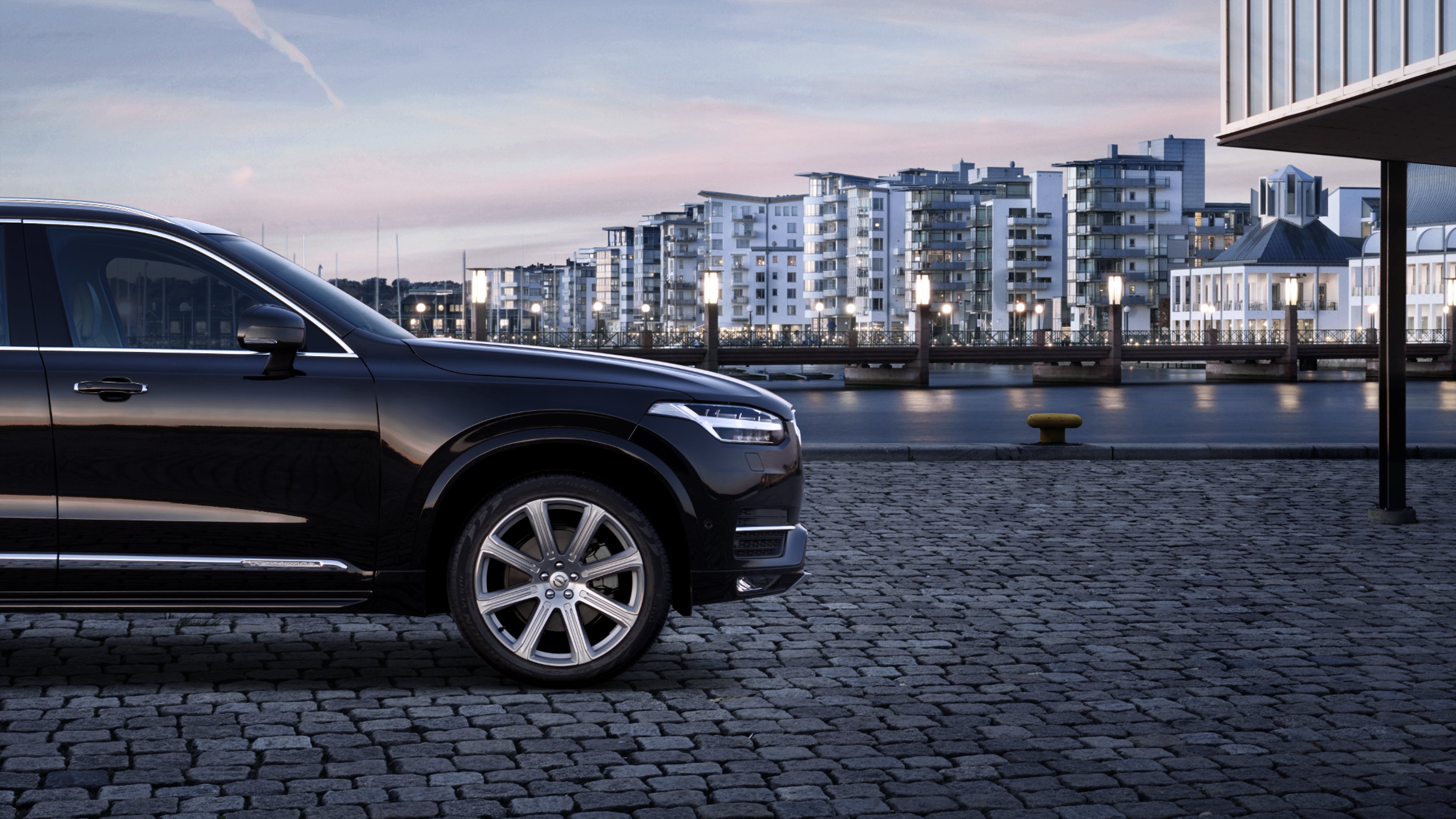 Vue latérale d’un Volvo XC90 noir garé sur une rue pavée, avec des immeubles modernes en arrière-plan