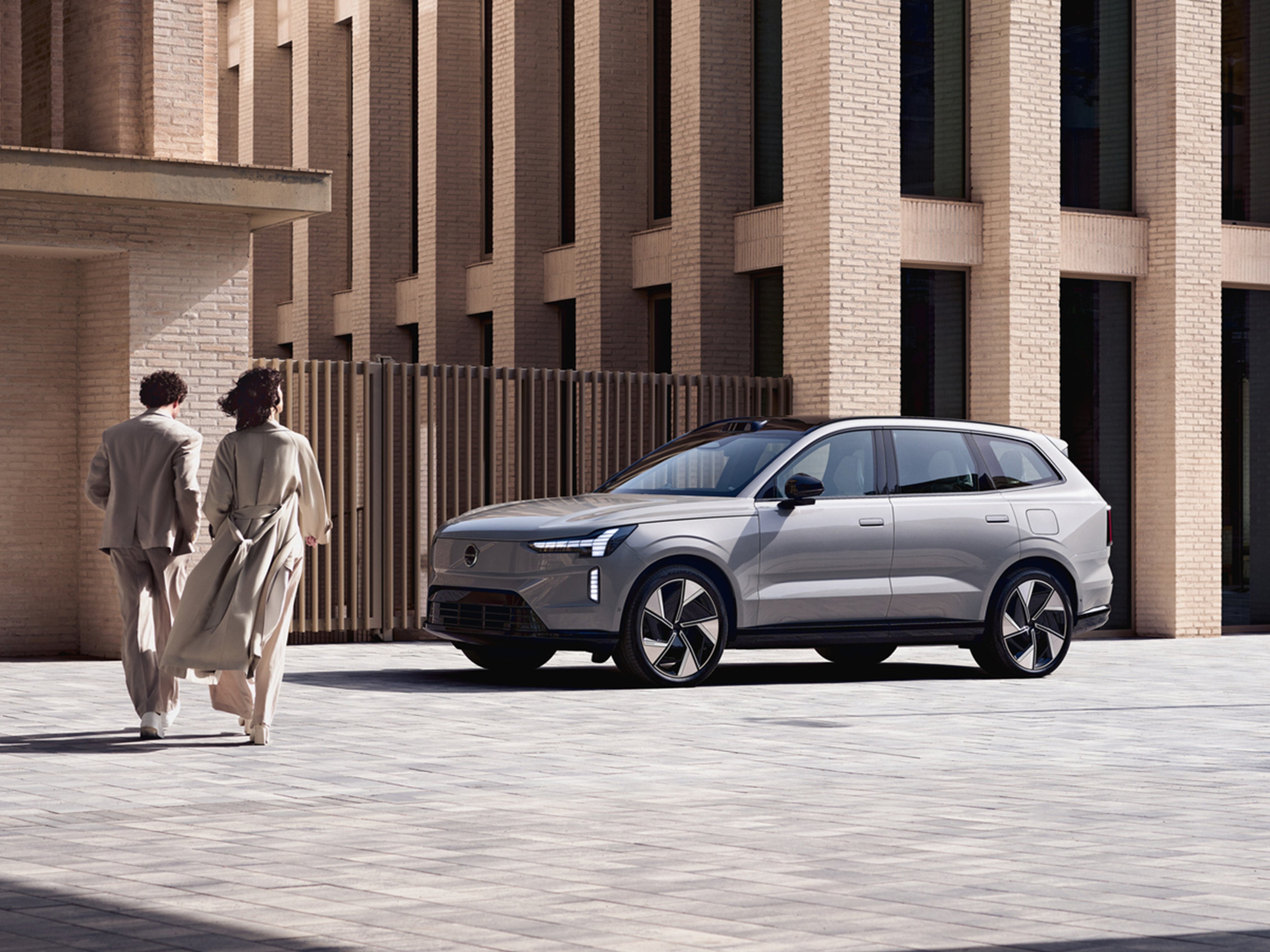 Nainen ja mies kävelee kohti Vapour Grey -väristä Volvo EX90 täyssähköautoa kaupungissa.