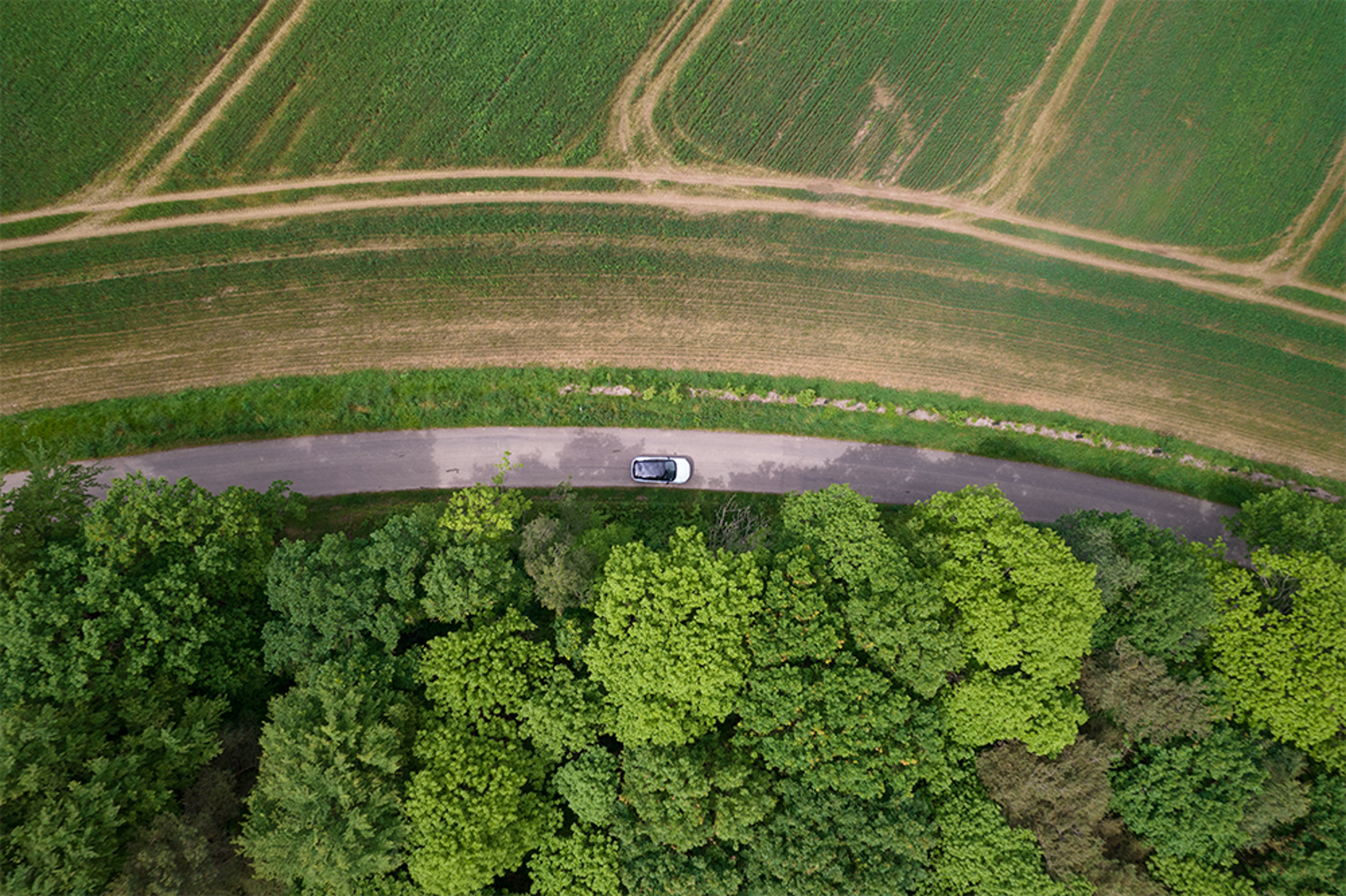 Vue aérienne d’une Volvo blanche sur route sinueuse entre champs verts et forêt dense.