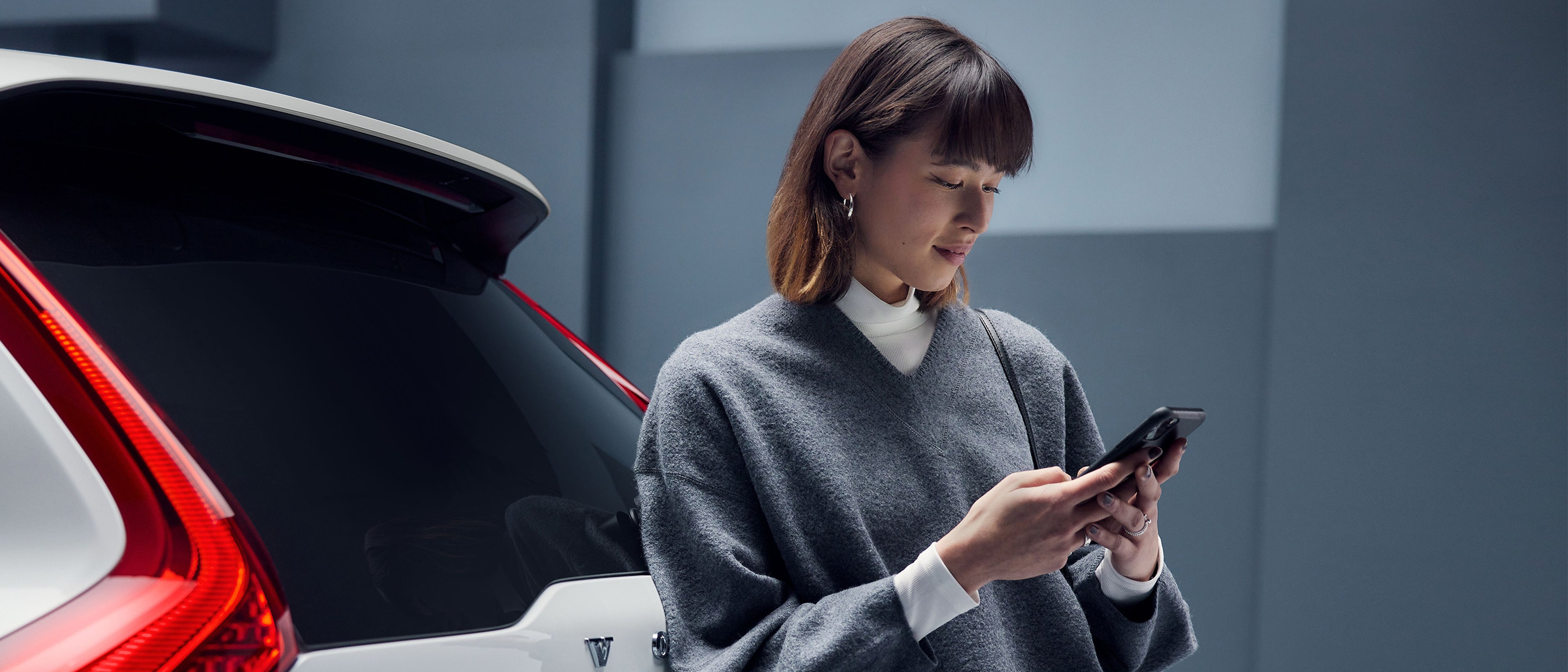 A woman wearing a gray sweater checks her phone in the Volvo app, standing beside a white Volvo in a modern indoor setting.