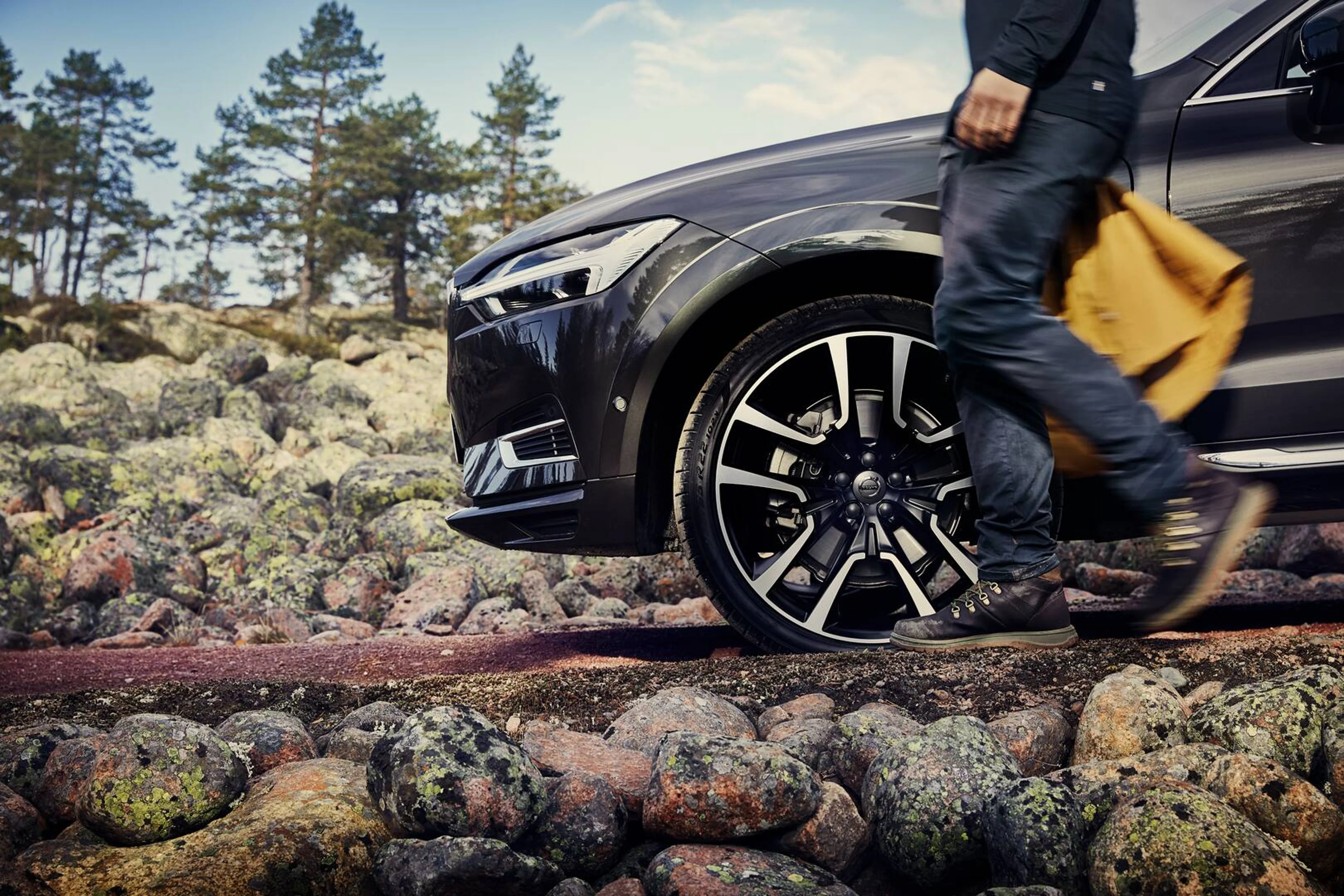 En person i jeans går forbi en sort Volvo bil og bærer en tanfarvet taske. Bilen er parkeret på klippefyldt terræn med sparsomme træer i baggrunden.