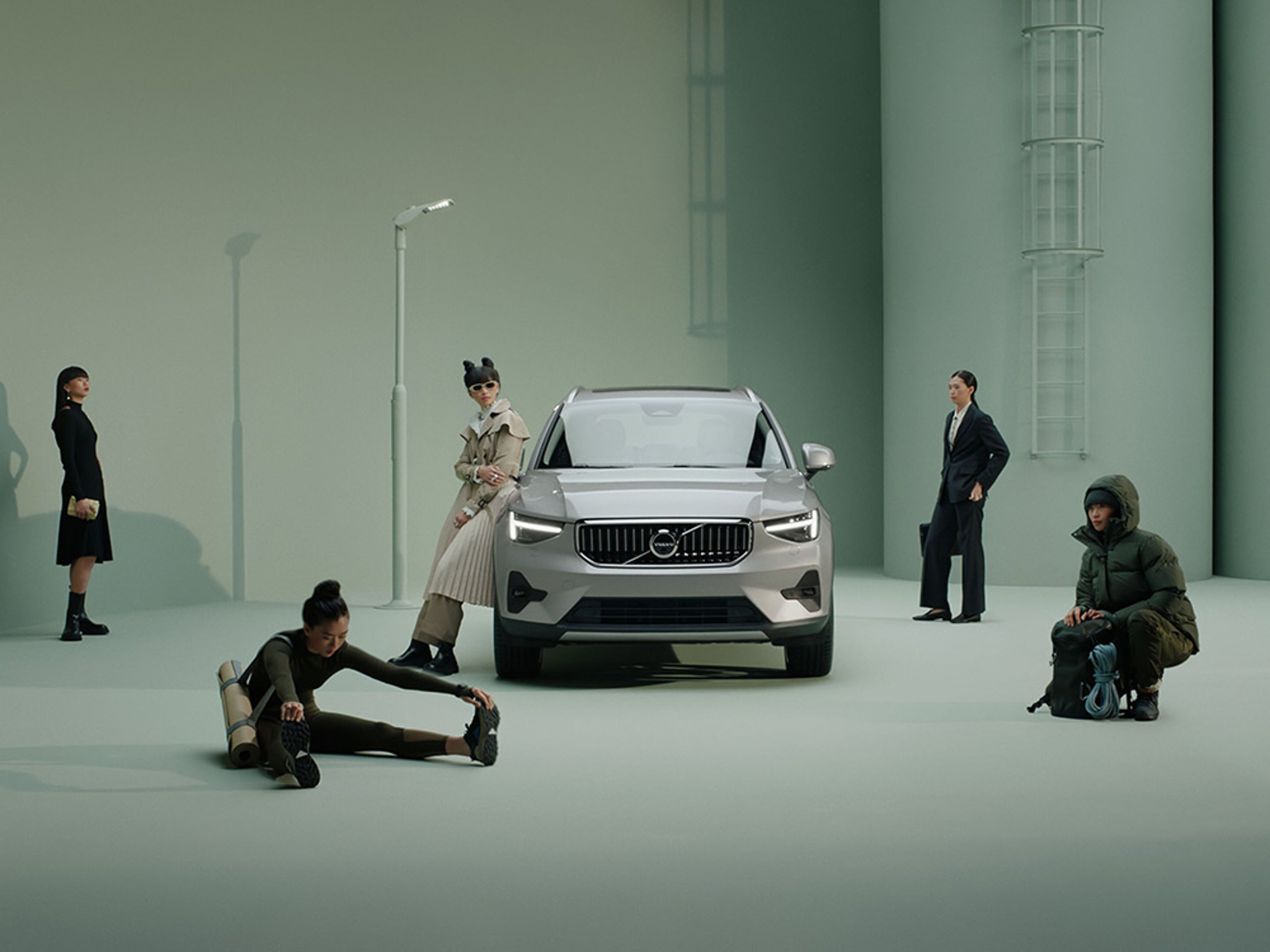 Diverse mensen in verschillende outfits staan rond een zilveren Volvo EX40 op een groene studio achtergrond.
